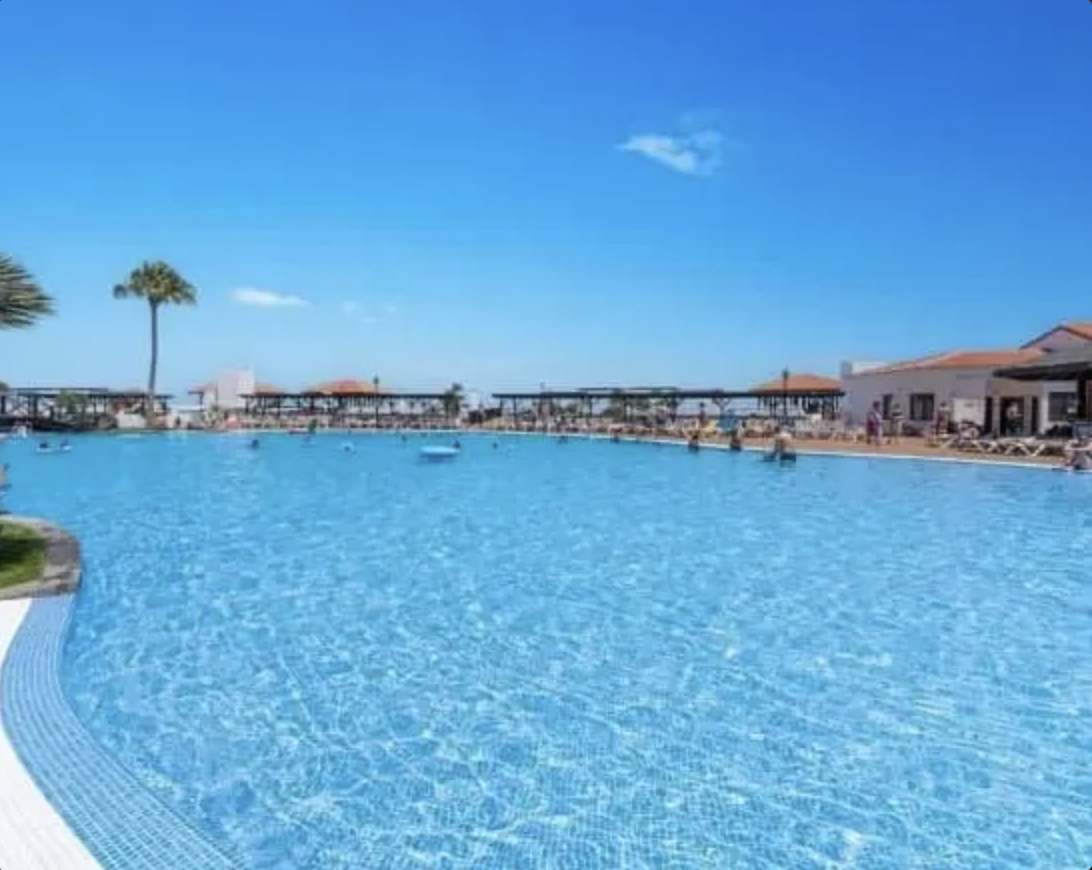 Piscina con acqua blu sotto un cielo azzurro, con alcune palme e strutture sullo sfondo.