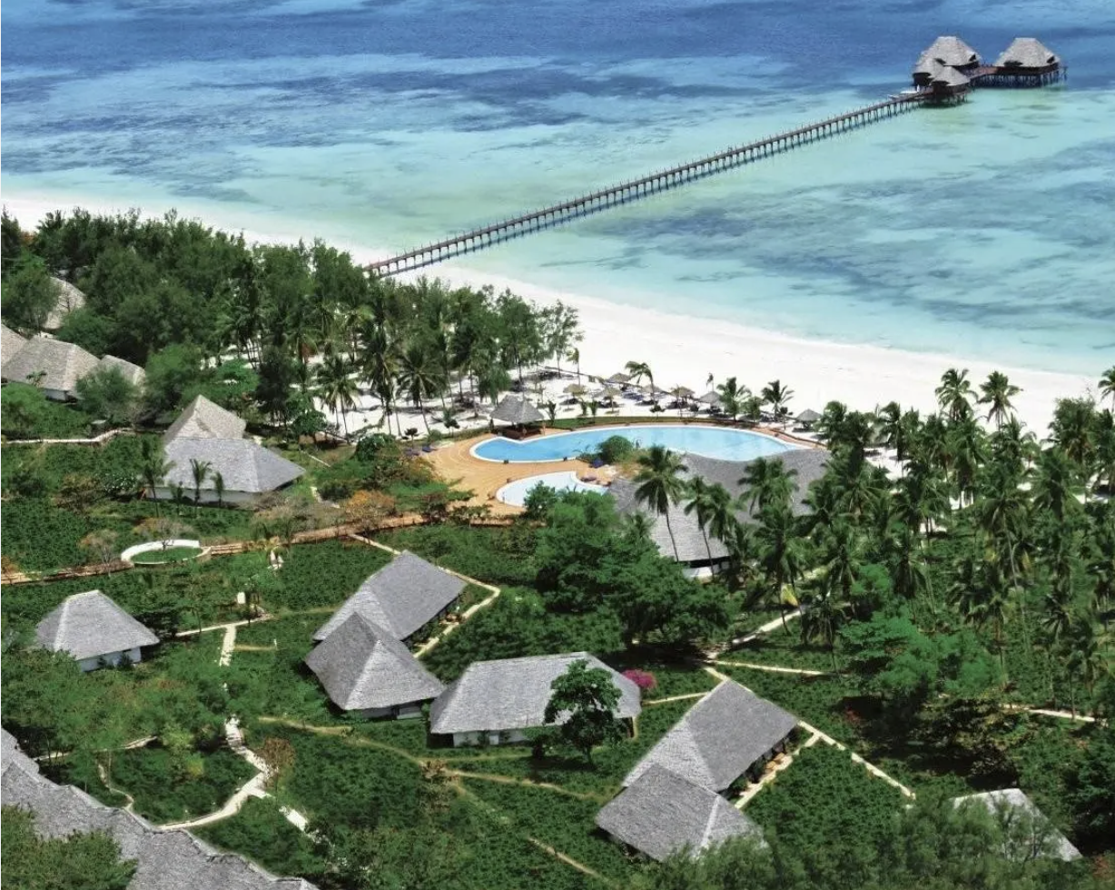Recensione aerea di un resort sulla spiaggia con piscine, bungalow di paglia, palme e un pontile che si estende nell'oceano.