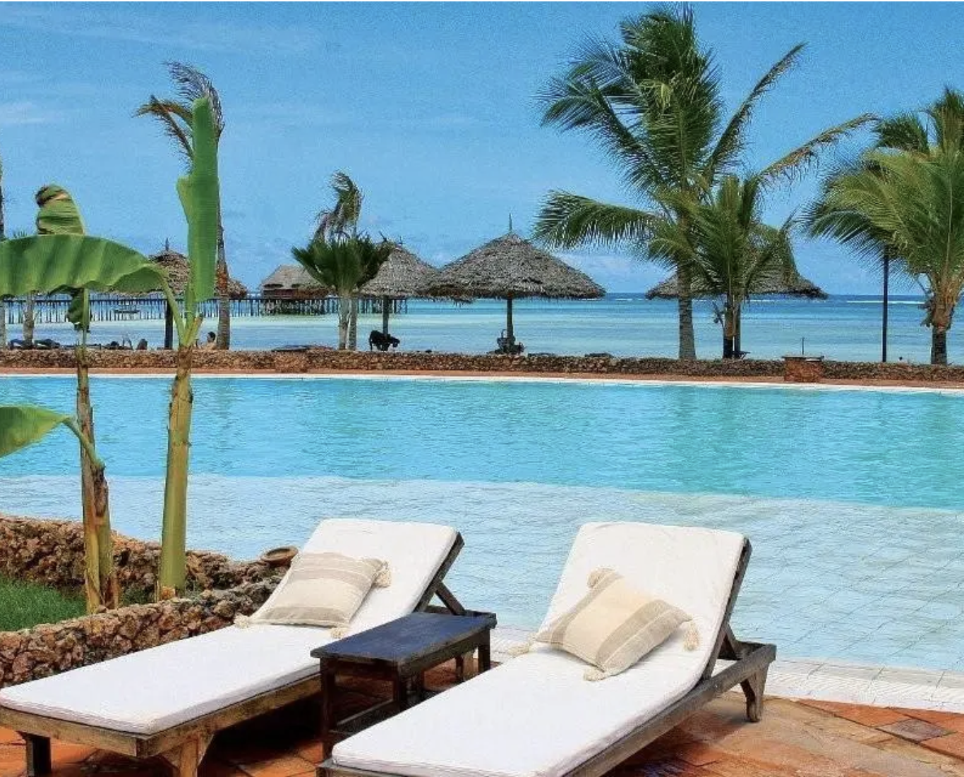 Due lettini con cuscini e un tavolino accanto, piscina con vista sull'oceano e palme in una spiaggia da sogno tropicale con capanne di paglia sullo sfondo.