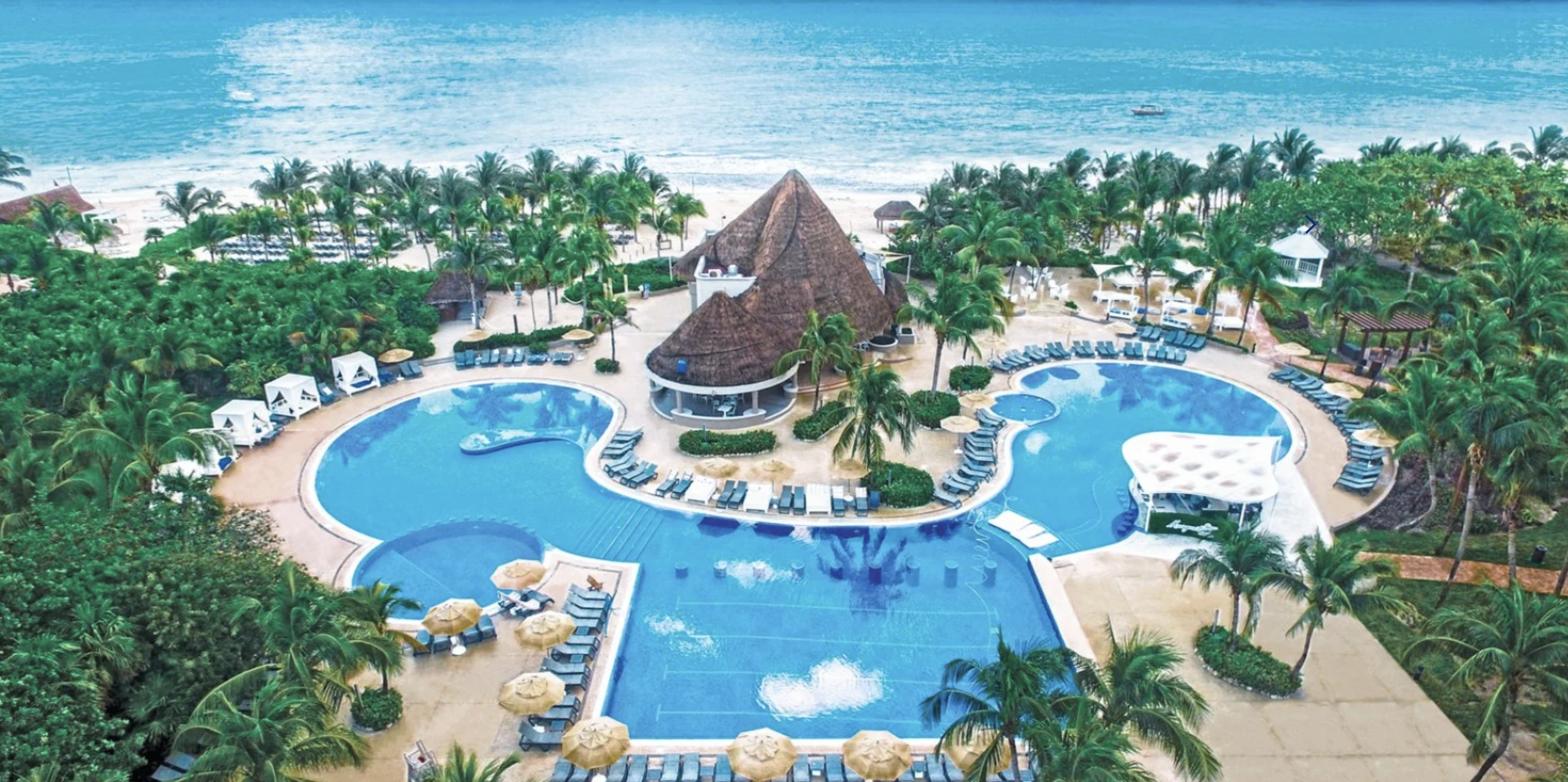 Vista aerea di una resort con piscine, palme e spiaggia con onde dell'oceano.