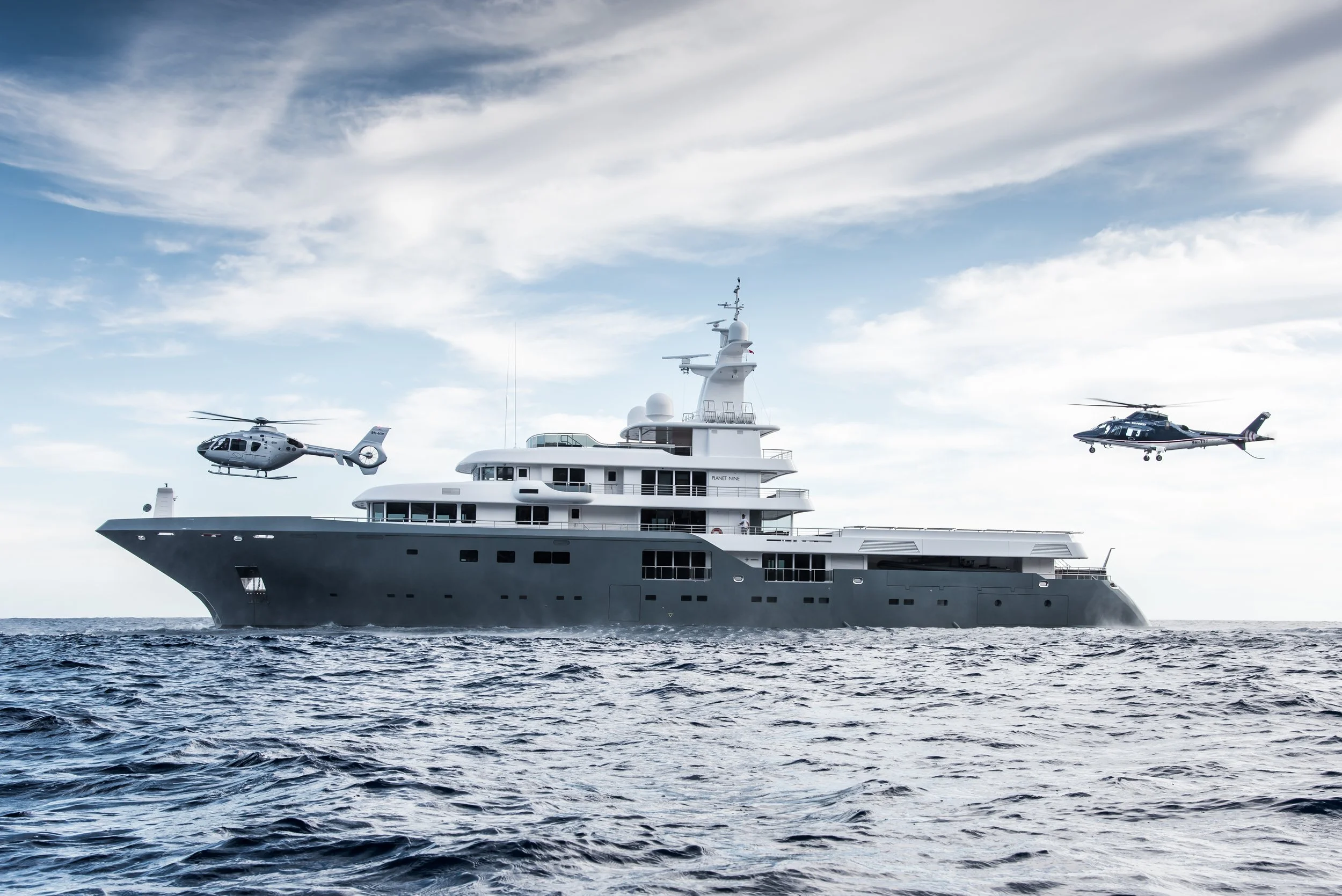 Large luxury yacht on the ocean with two helicopters flying nearby, under partly cloudy sky.
