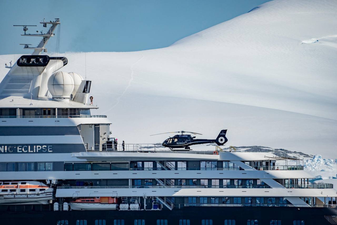 A luxury cruise ship named 'NICE ECLIPSE' in a snowy, icy landscape with a helicopter on the deck and snow-covered mountains in the background.