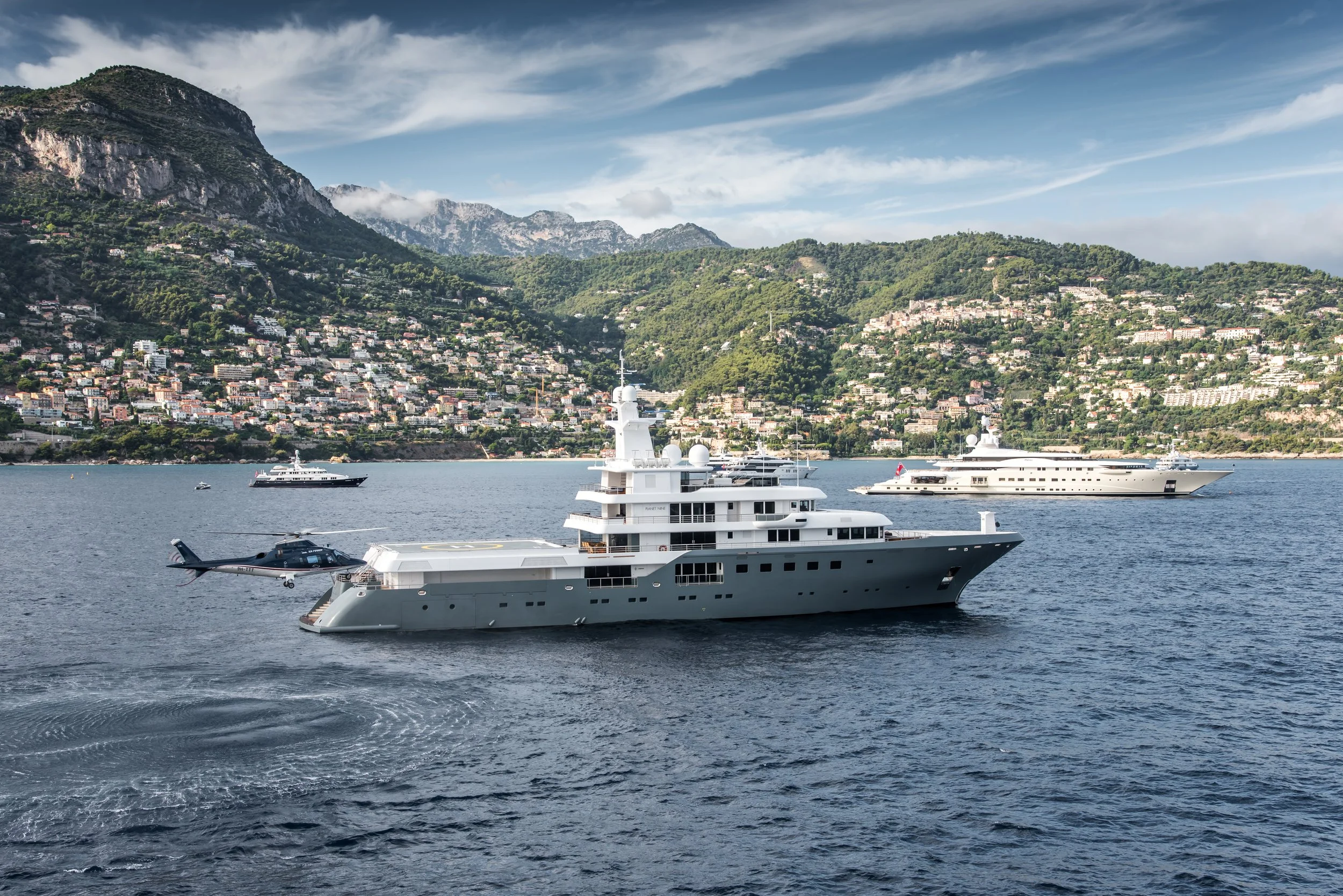 Multiple luxury yachts anchored in a bay with green mountains and a town in the background, under a partly cloudy sky.