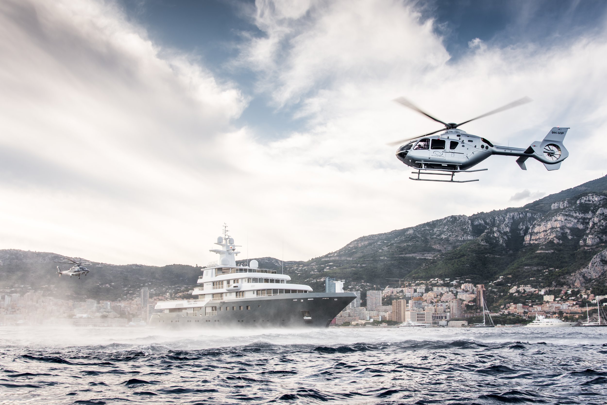 A large luxury yacht cruising on the water with two helicopters flying overhead against a backdrop of mountains and a cityscape.