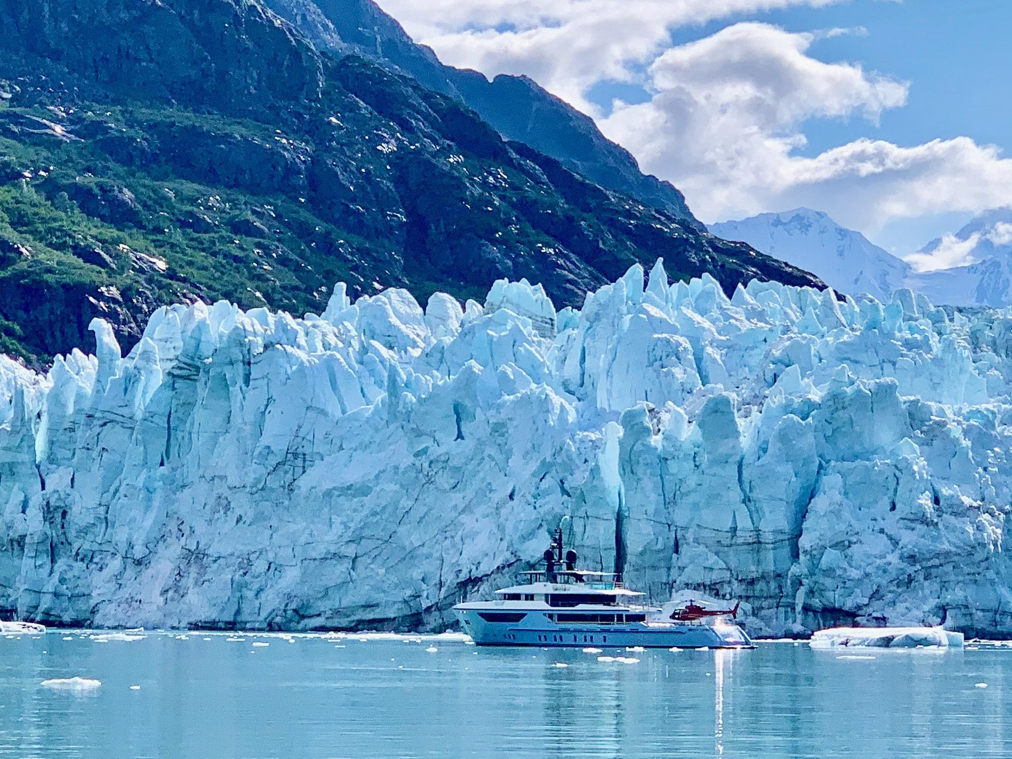 A yacht sailing near large glacial ice formations with mountain and cloudy sky in the background.