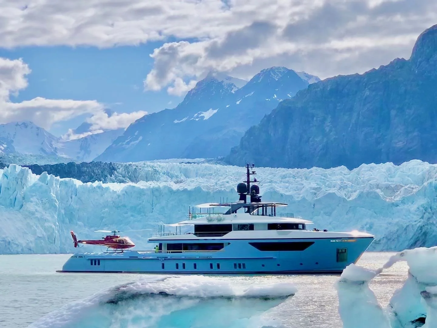 A luxury yacht with a red helicopter is sailing on icy waters near a glacier, with snow-covered mountains and a cloudy sky in the background.