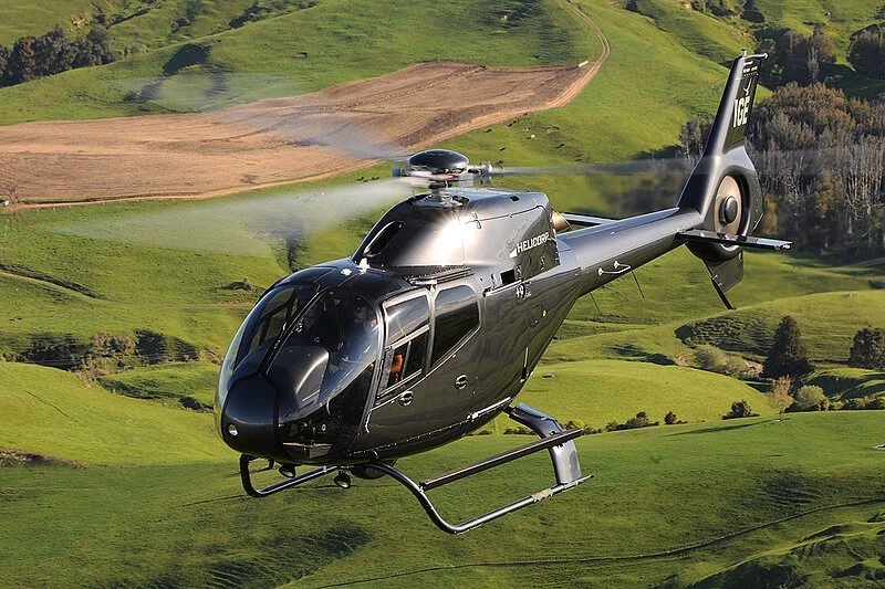 A black helicopter flying over green rolling hills and farmland.
