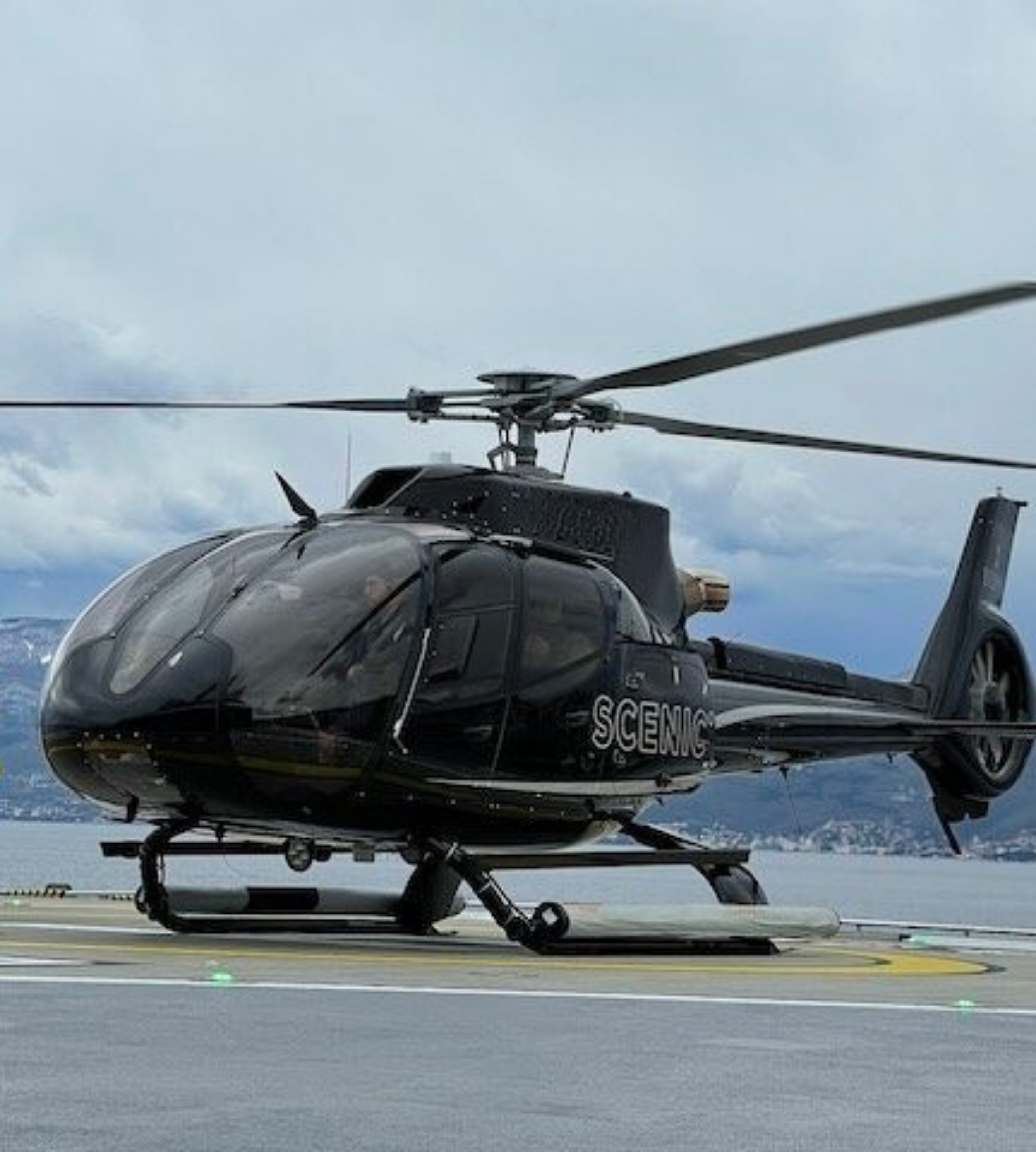 Black helicopter on a helipad with mountains and cloudy sky in background.