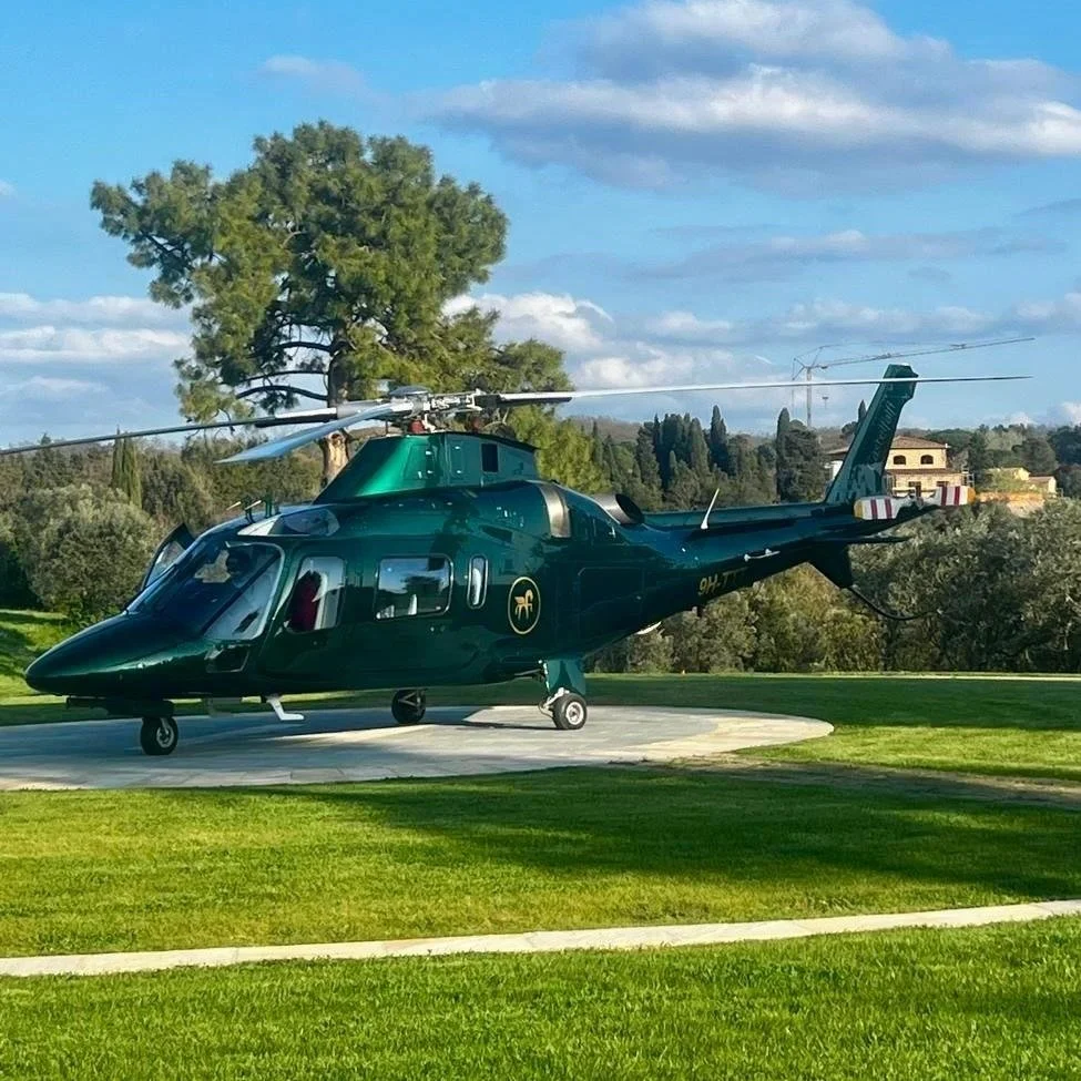 A dark green helicopter on a circular landing pad surrounded by grass, with trees and buildings in the background, and a cloudy sky overhead.
