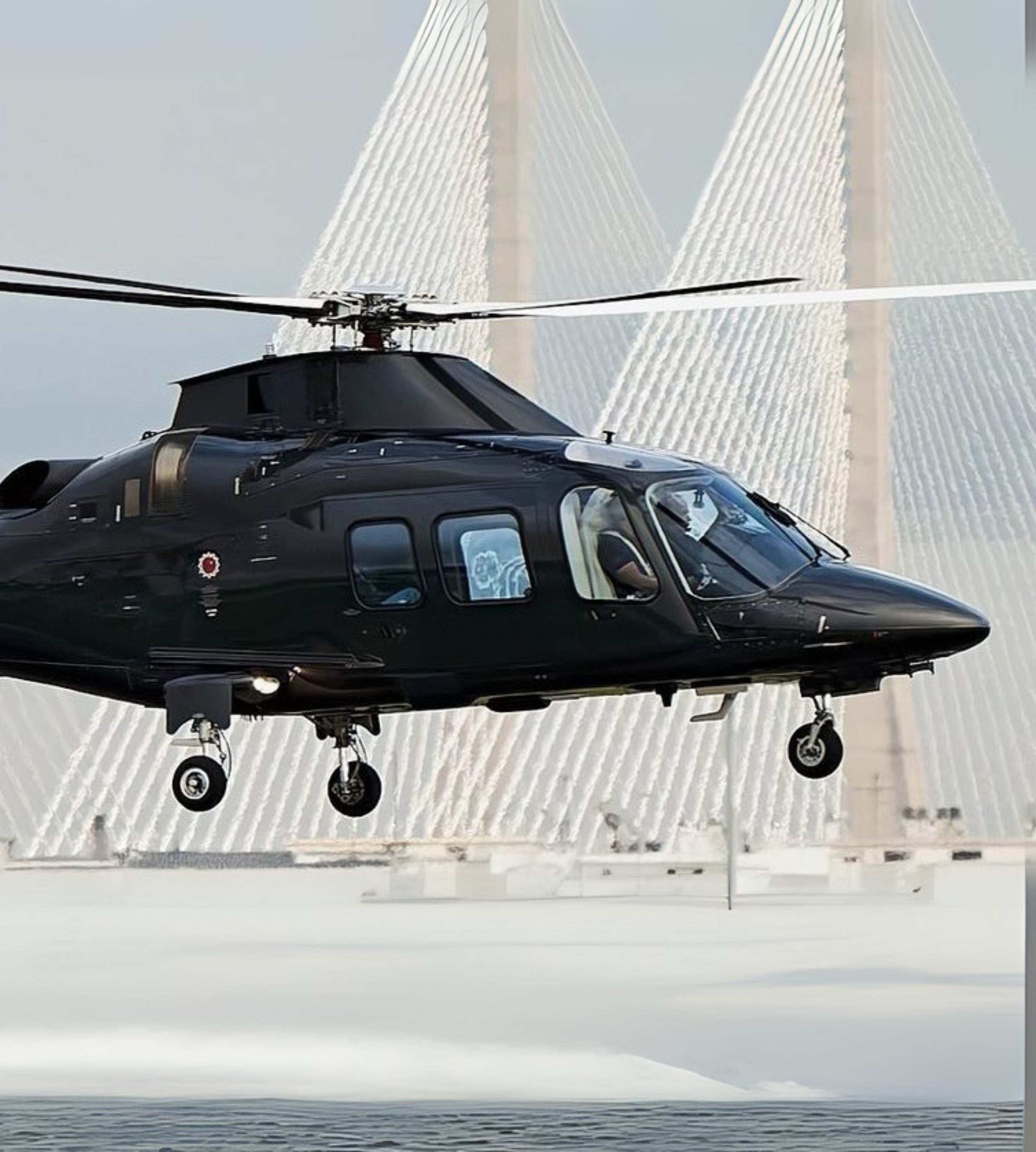 Black helicopter flying near a large cable-stayed bridge with white cables and a tall tower.