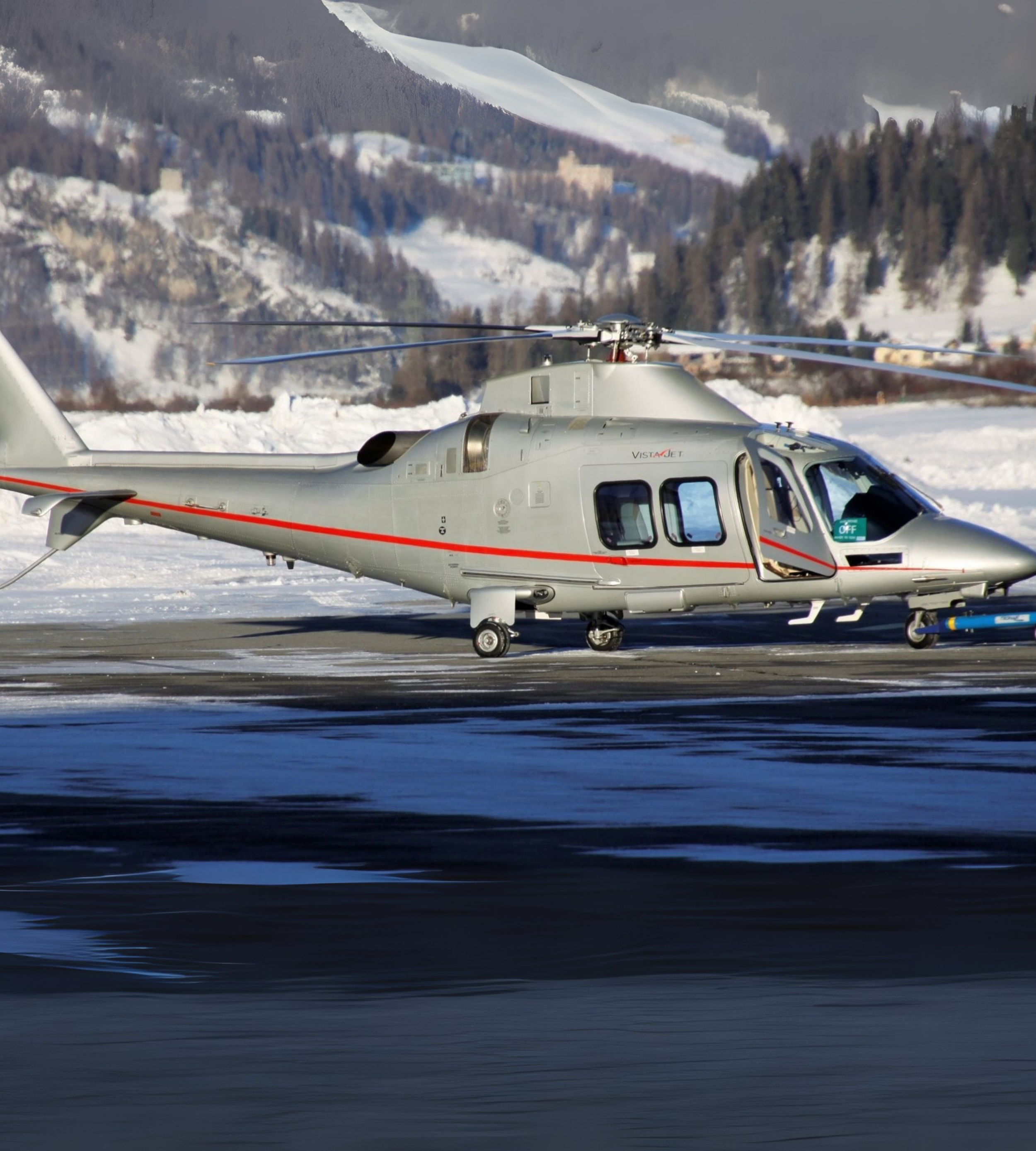 A gray helicopter on a snowy tarmac with a mountain landscape in the background.