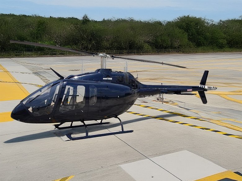 A black helicopter parked on a concrete helipad with yellow markings, surrounded by greenery and a partly cloudy sky.