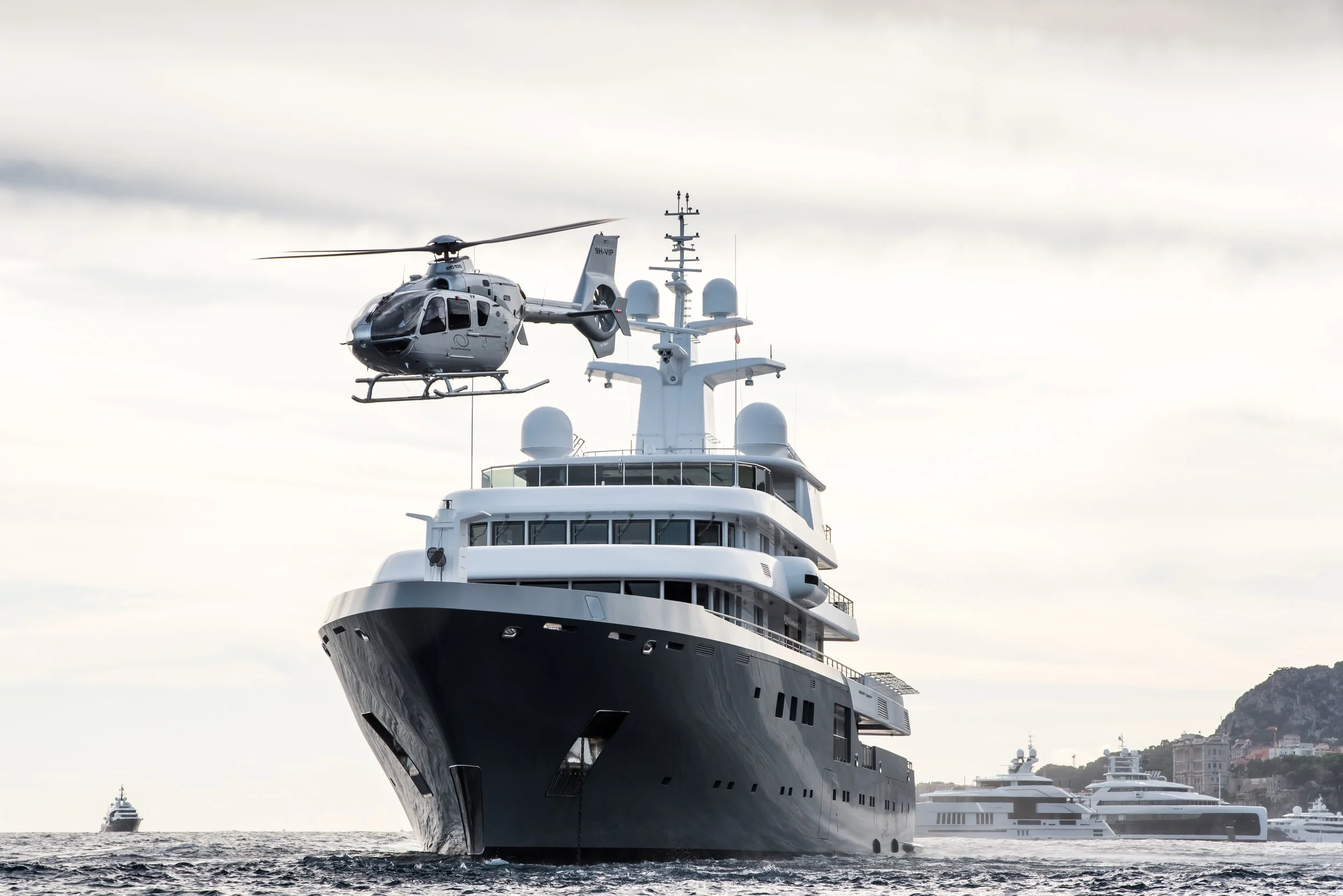 Large luxury yacht with a helicopter flying above it in the ocean, with other yachts and a hill in the background.