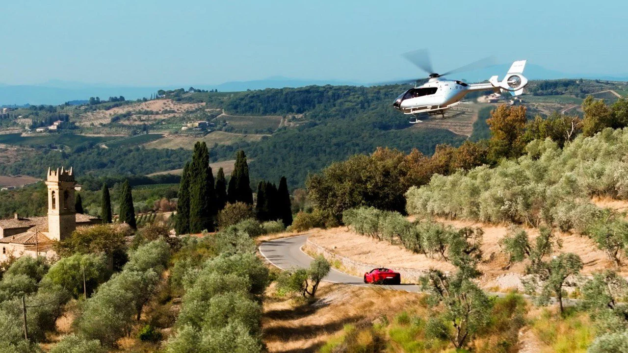 A helicopter flying over a scenic hillside with green trees, olive groves, a winding road, and a red sports car.
