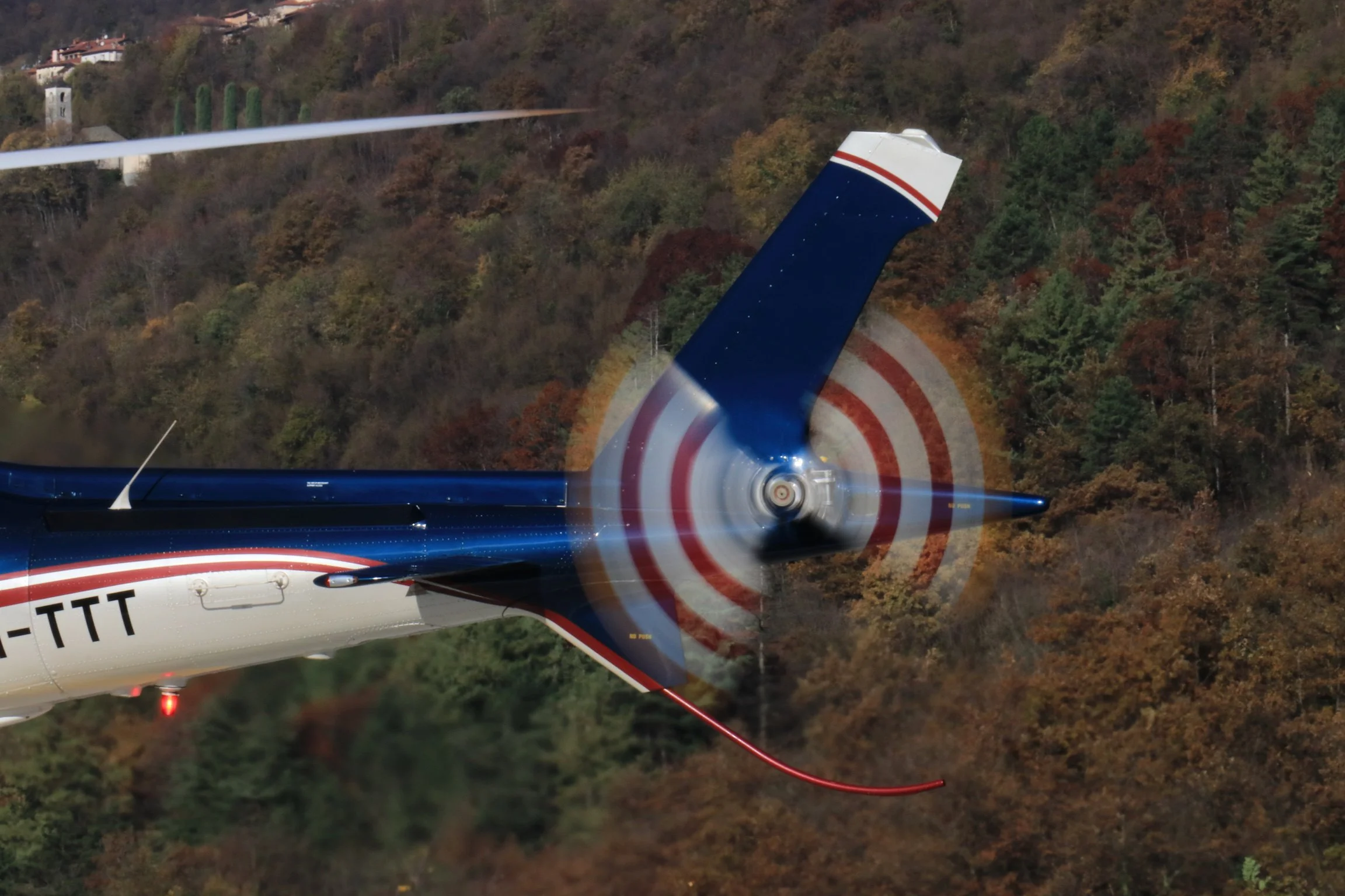 A helicopter with spinning rotor blades flying over a wooded area with autumn foliage.