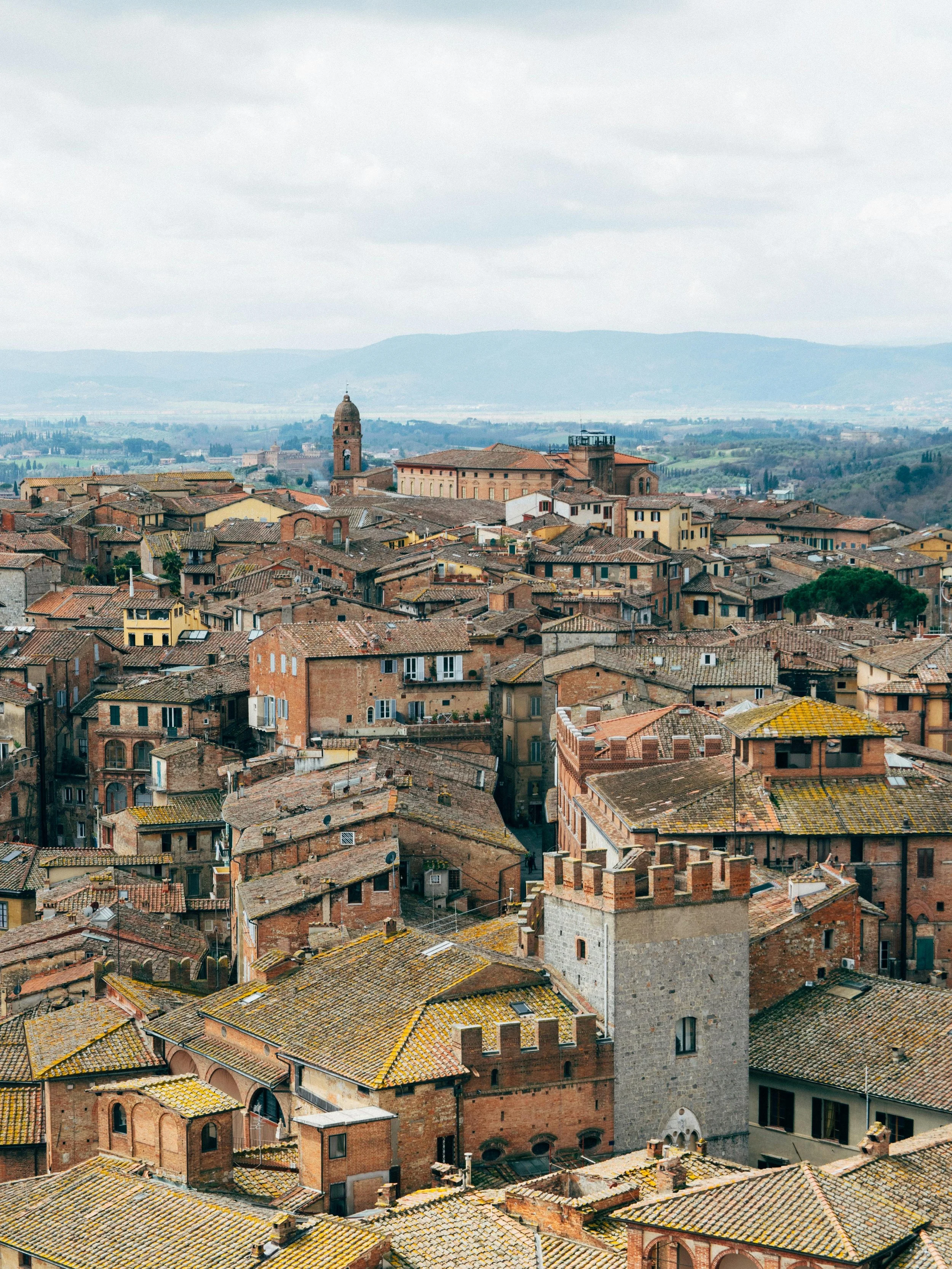 Aerial view of an old European city with red-tiled roofs, stone towers, and narrow streets, surrounded by rolling hills and distant mountains.