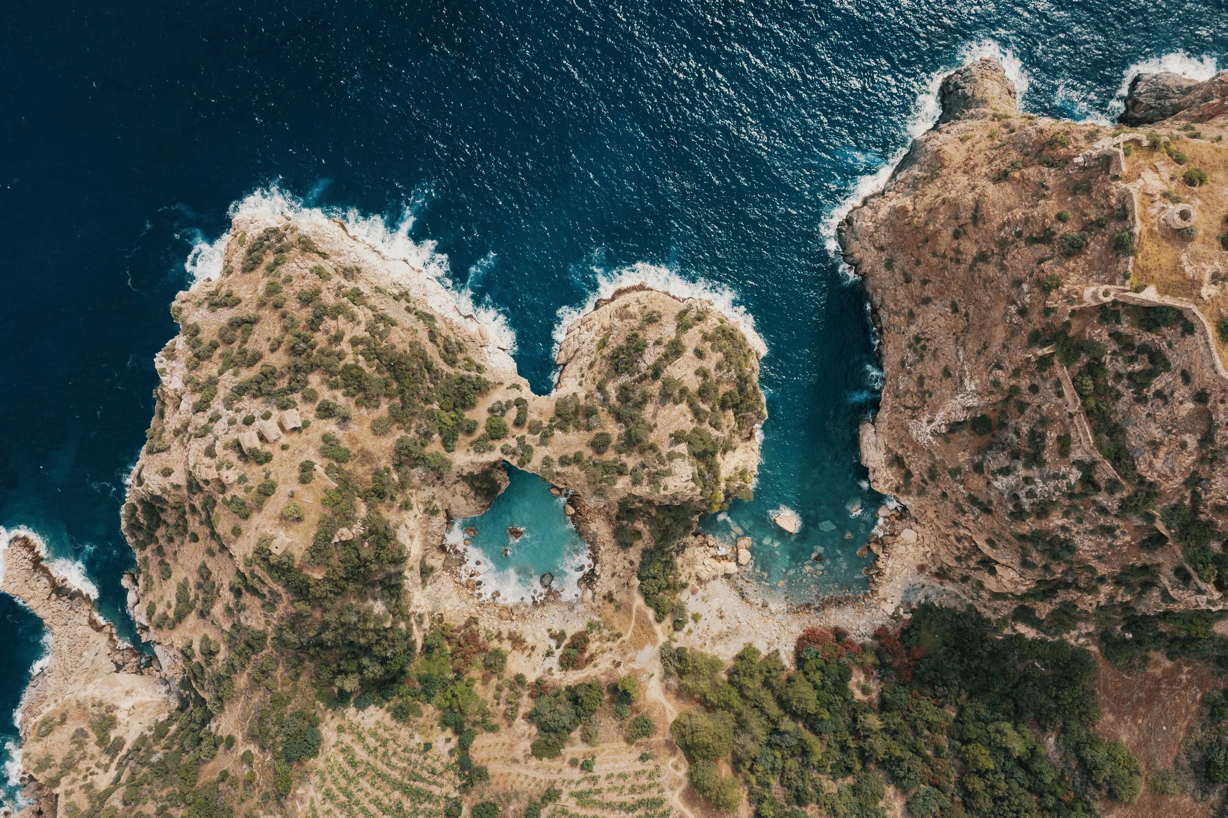 Aerial view of rugged coastline with two large rocky formations in the sea, surrounded by greenery and clear blue waters.