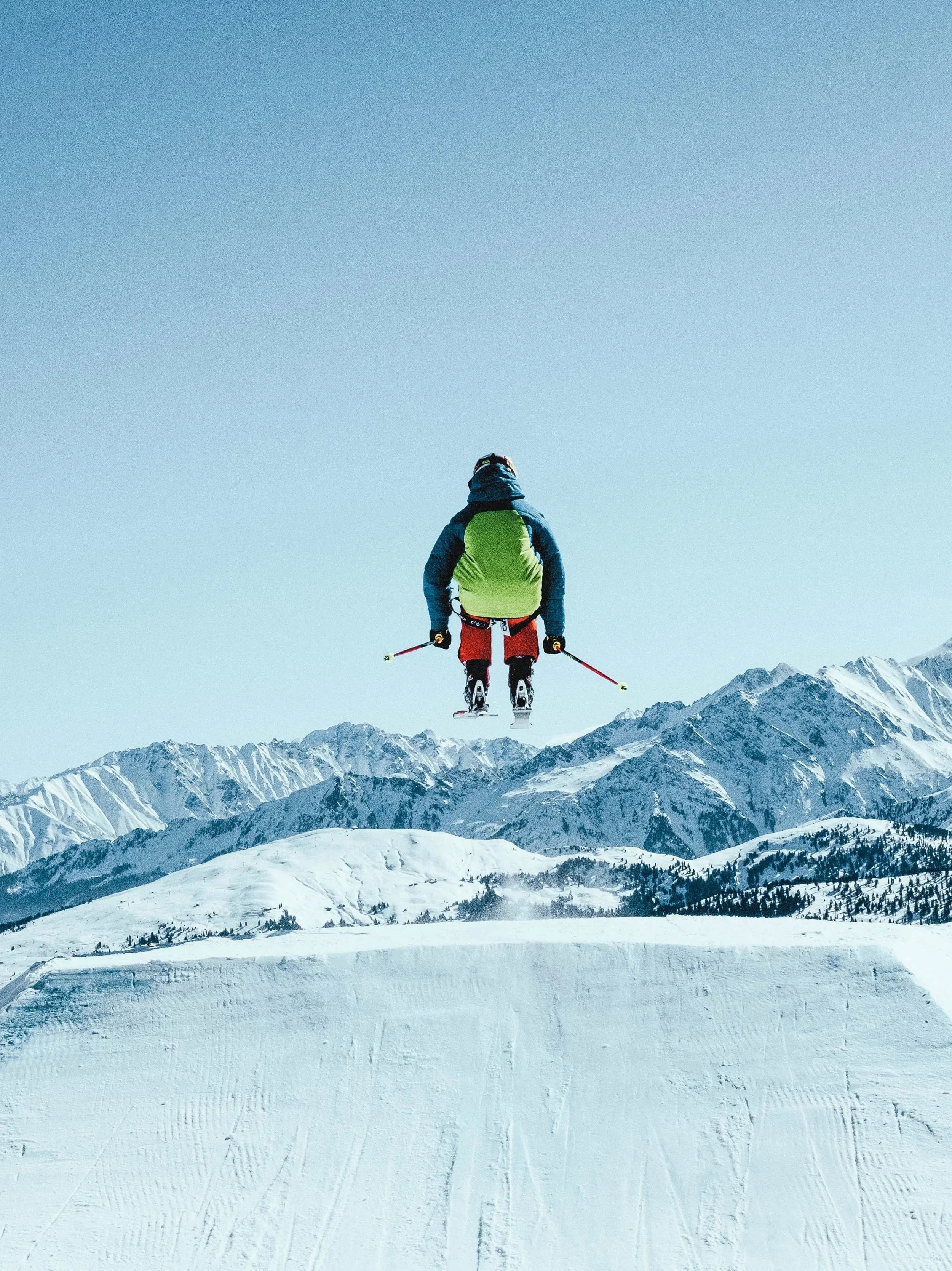 Skier jumping off a snow ramp in a snowy mountain landscape with clear blue sky.