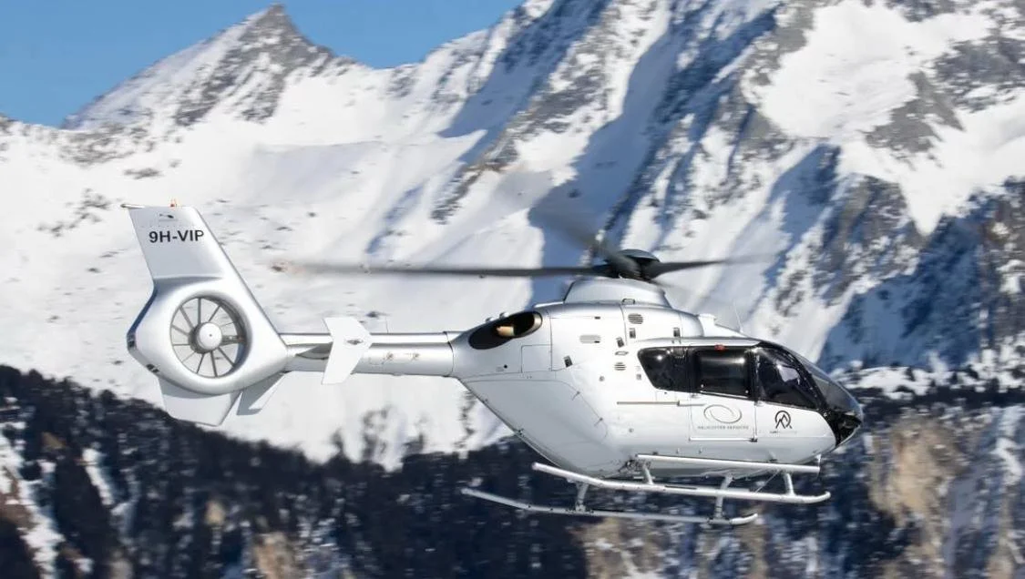 White helicopter flying in front of snow-covered mountains and blue sky.