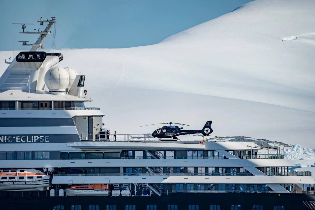 The image shows a luxury cruise ship named 'NICE ECLIPSE' docked in an icy, snow-covered area. There is a helicopter parked on the top deck of the cruise ship, and the background features a snow-covered landscape.