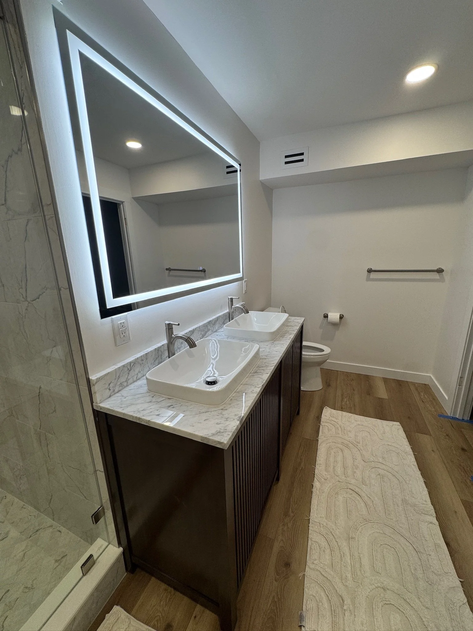 Modern bathroom with a dual vanity featuring two rectangular vessel sinks and a large backlit mirror. There is a wooden cabinet under the marble countertop, a toilet, a towel rack, a bathmat, and part of a glass shower with marble tiling visible on t