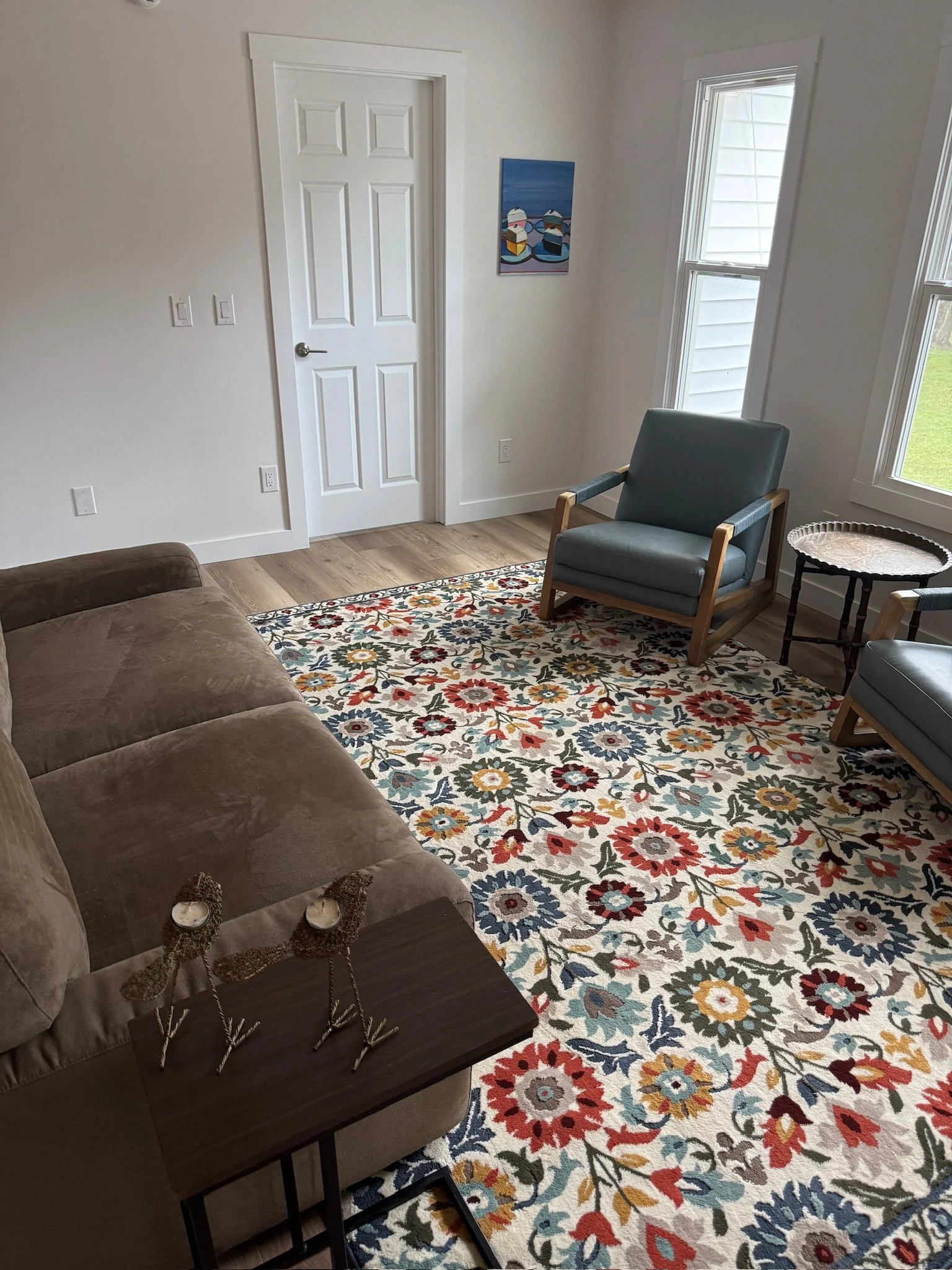 Living room with a brown sofa, two teal armchairs, a floral patterned area rug, a small wooden side table, and artwork on the wall.
