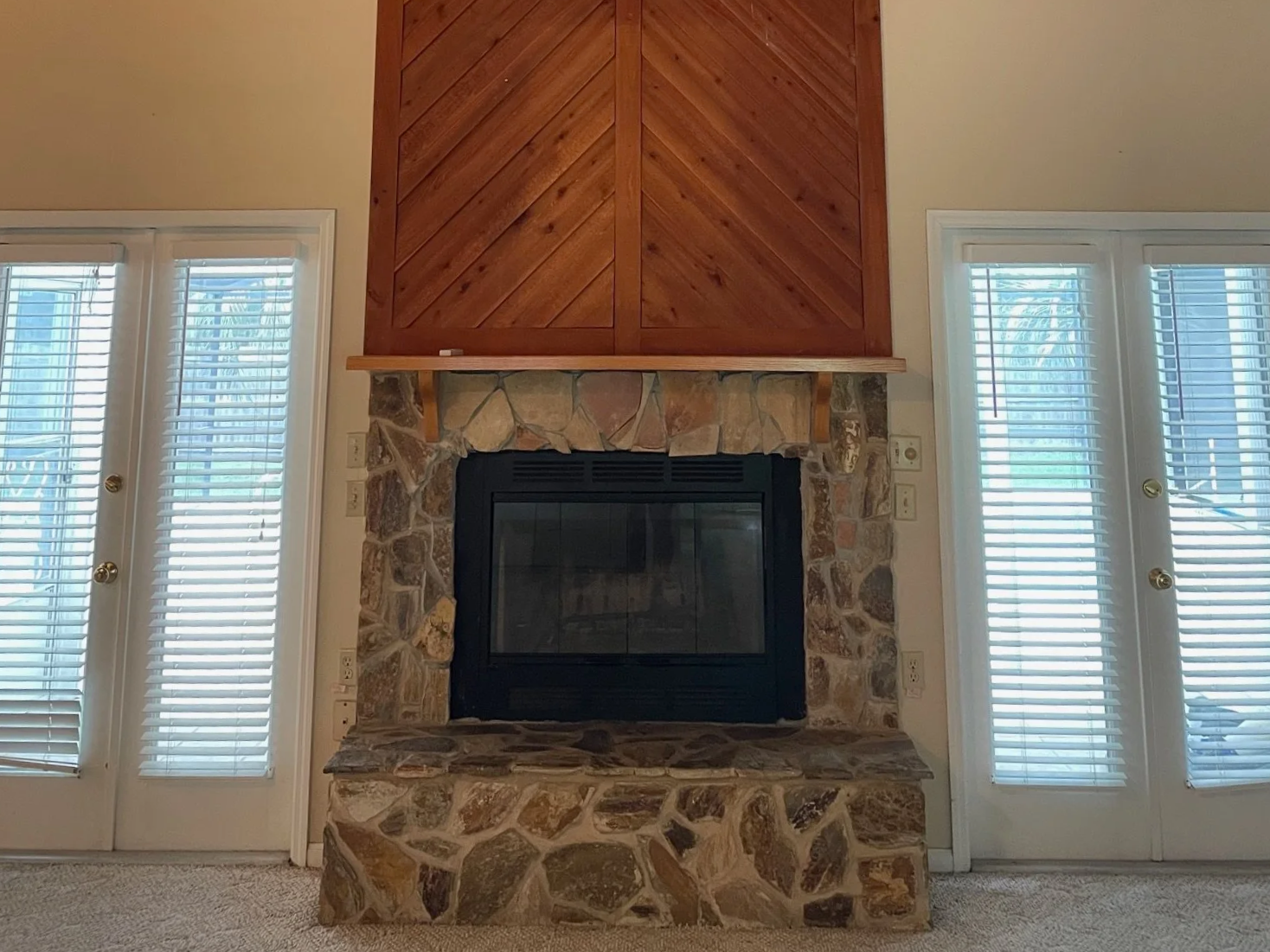 Living room with a stone fireplace, wooden mantle, and wood-paneled wall above fireplace, flanked by glass doors with blinds.