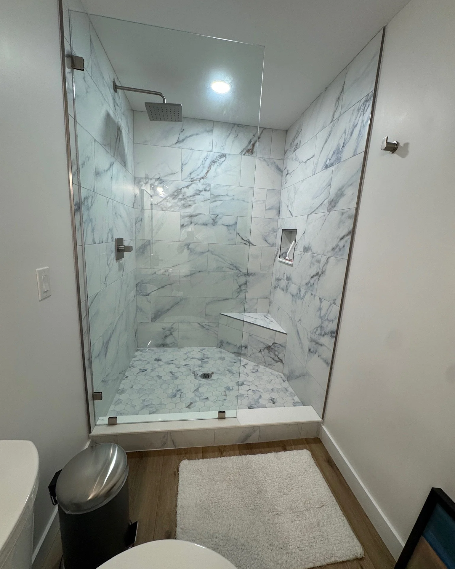 Bathroom with a glass-enclosed marble shower featuring a built-in bench, a wall niche, and rainfall showerhead, with a white rug on wooden floor in front.