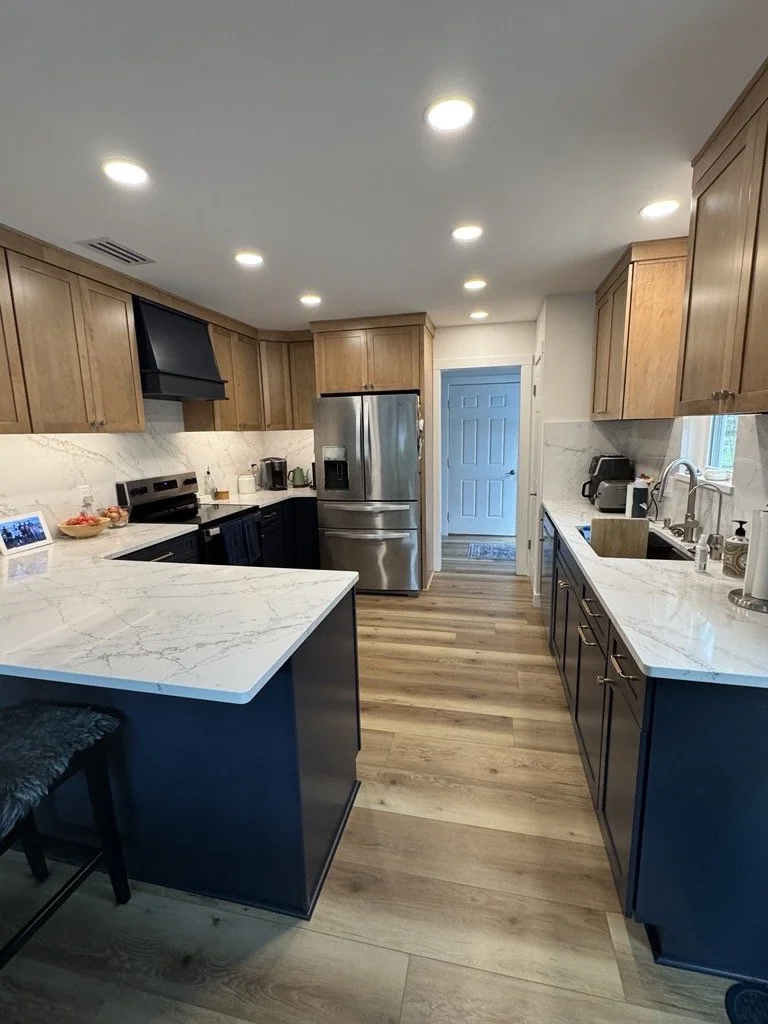 Modern kitchen with wooden cabinets, white marble countertops, stainless steel appliances, blue lower cabinets, and wooden flooring.