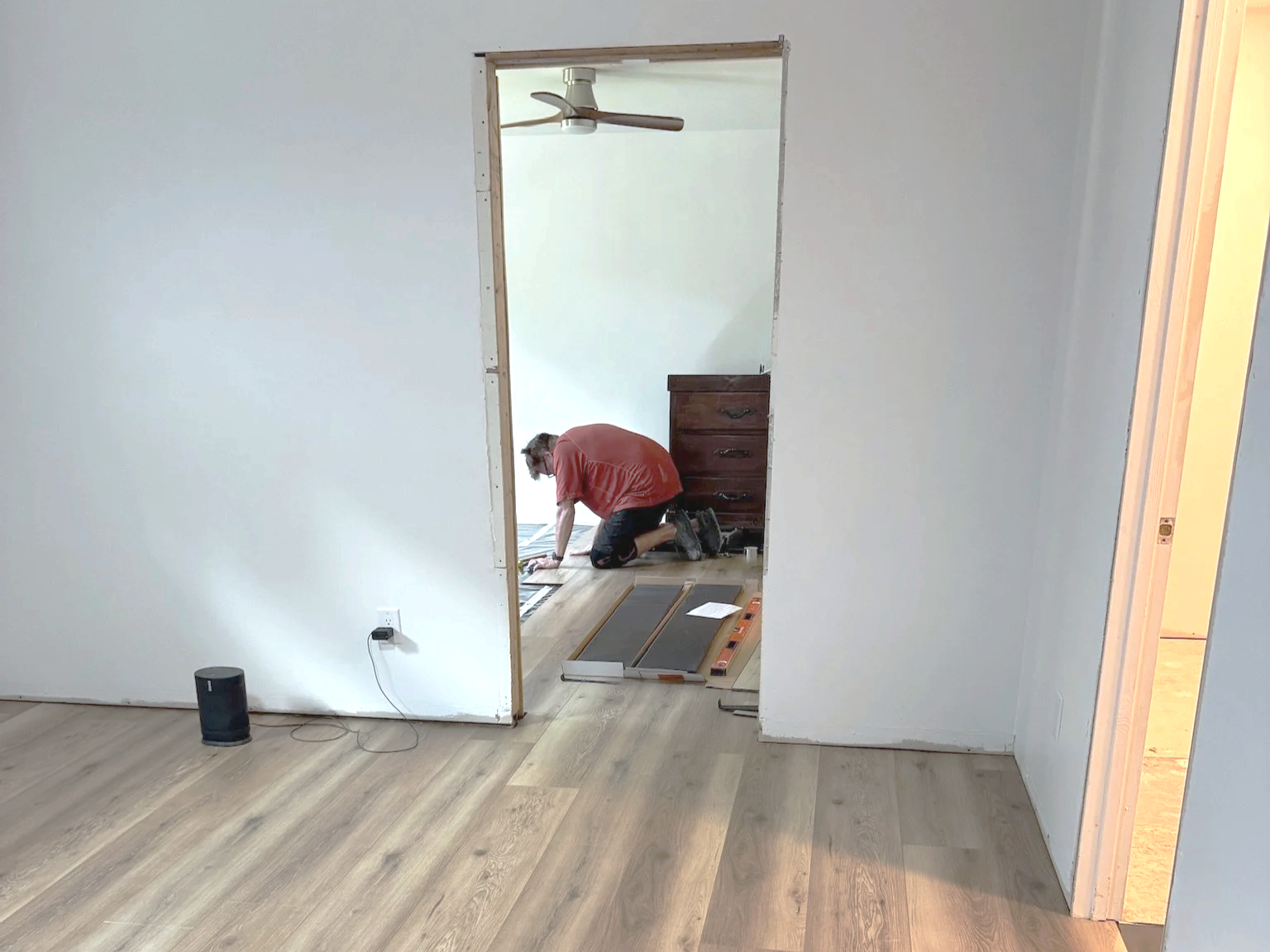A person kneeling on the floor working on installing or repairing wood flooring in a room. The room has white walls, a wooden floor, and an unfinished doorway leading to another room with a dresser and a ceiling fan.