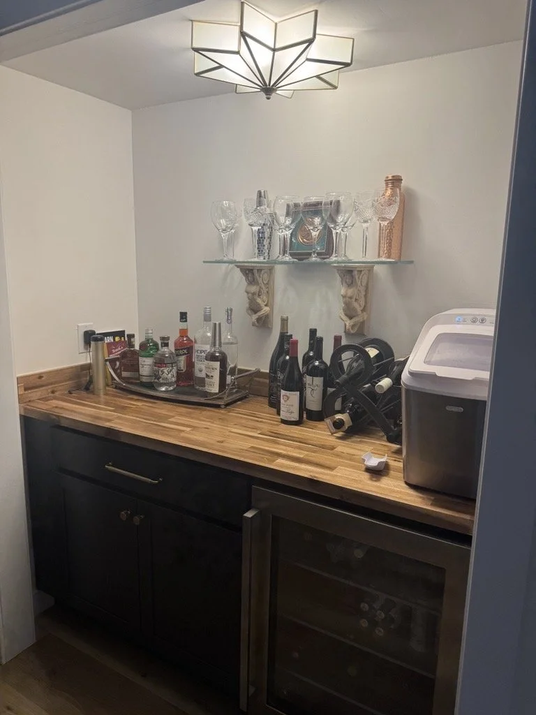 Bar area with a wooden countertop, wine bottles, liquor bottles, wine glasses, glass shelf with additional glasses and decorative items, wine chiller, and a modern ceiling light fixture.