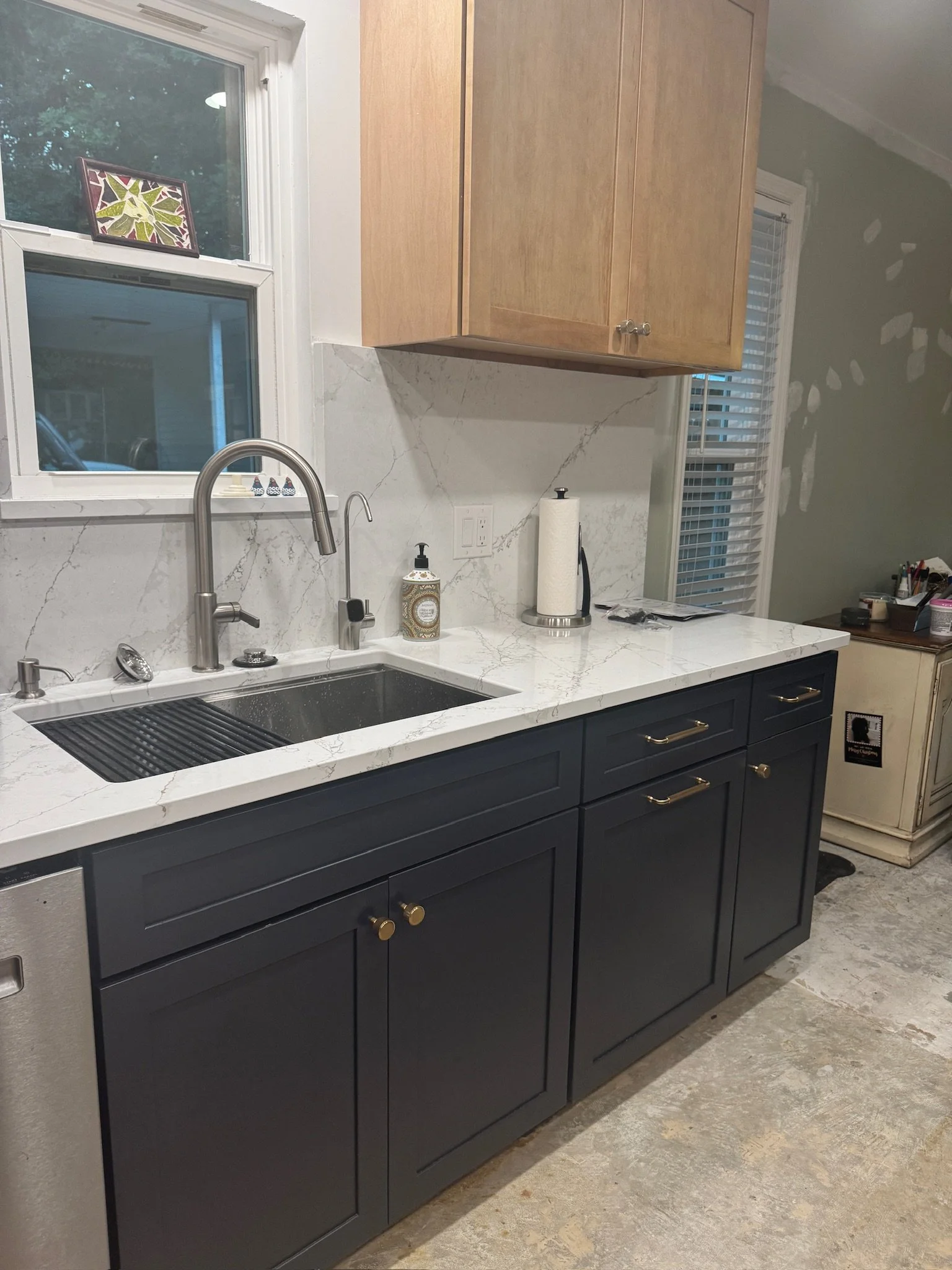 Kitchen with black lower cabinets, light wood upper cabinets, a white marble countertop and backsplash, a window above the sink, and various kitchen items on the counter.