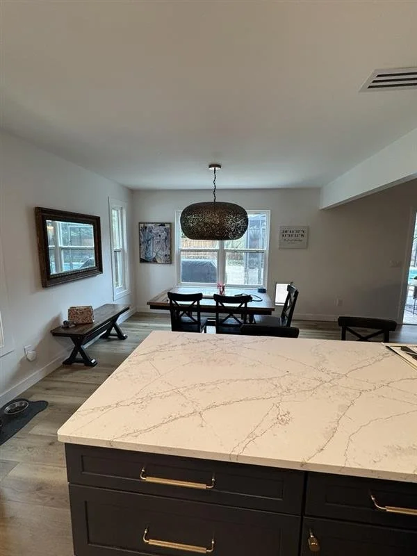 View of a dining area with a marble-topped island in the foreground, a wooden dining table with chairs, a pendant light, and windows in the background in a modern home interior.