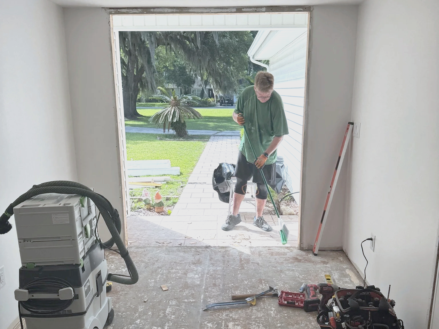 Man cleaning a patio outside a house under construction with tools and equipment inside the room.