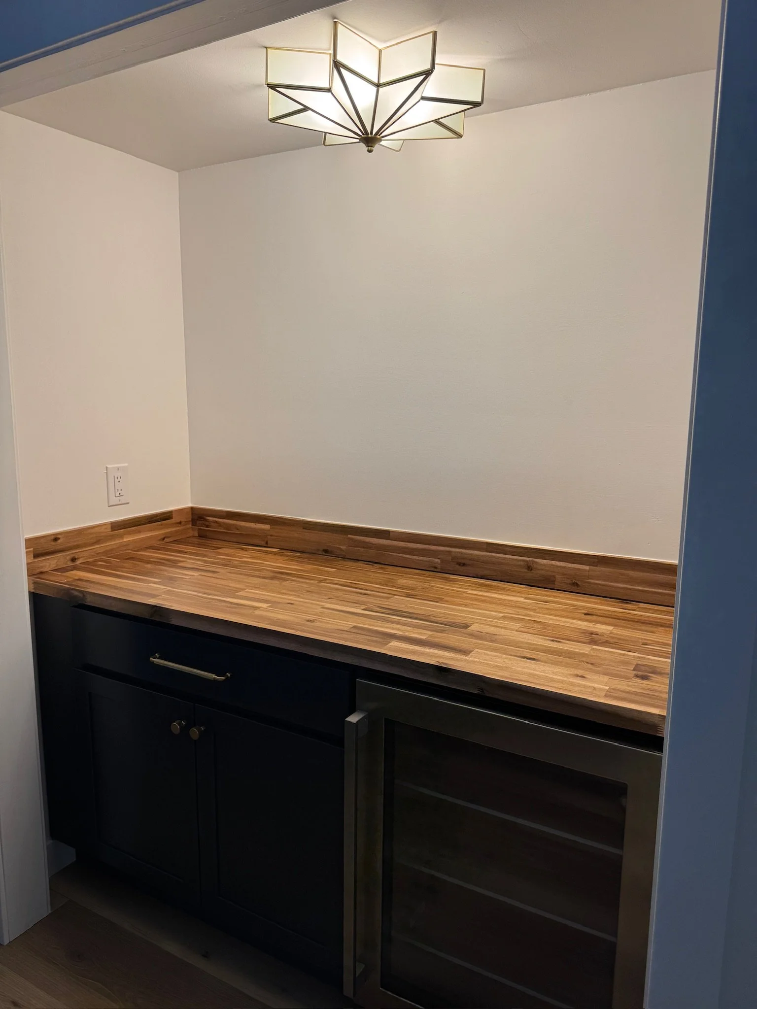 A small bar area with a wooden countertop, black cabinet, built-in wine cooler, and modern geometric ceiling light fixture.