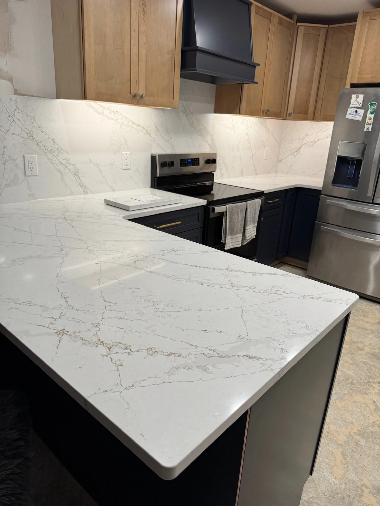 Modern kitchen with white marble countertops and backsplash, wooden upper cabinets, dark lower cabinets, stainless steel refrigerator, and black electric stove with a range hood.
