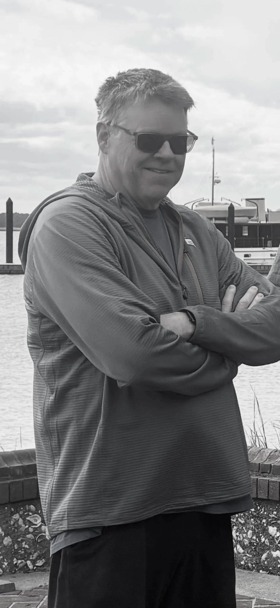 A man in sunglasses and a light zip-up jacket standing outdoors near water, with boats and a cloudy sky in the background.