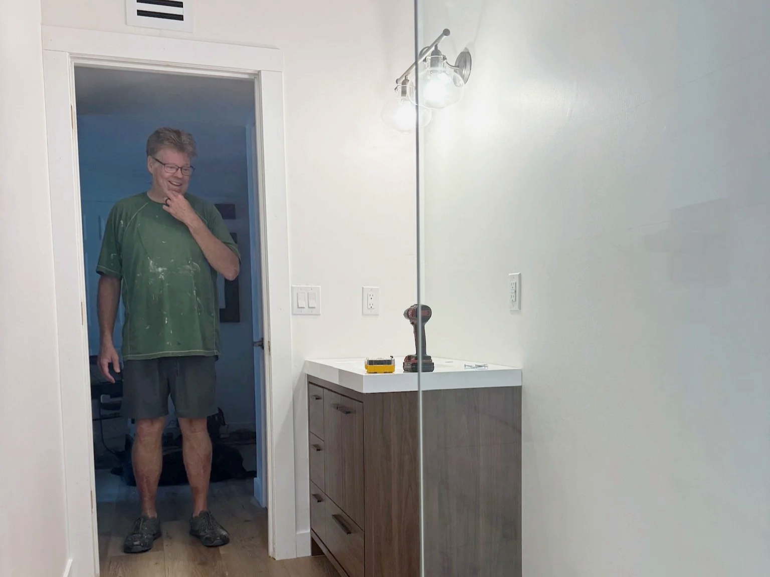 A man standing in front of a mirror with his reflection visible, wearing a green t-shirt and shorts, with tools and a drill on the bathroom counter.