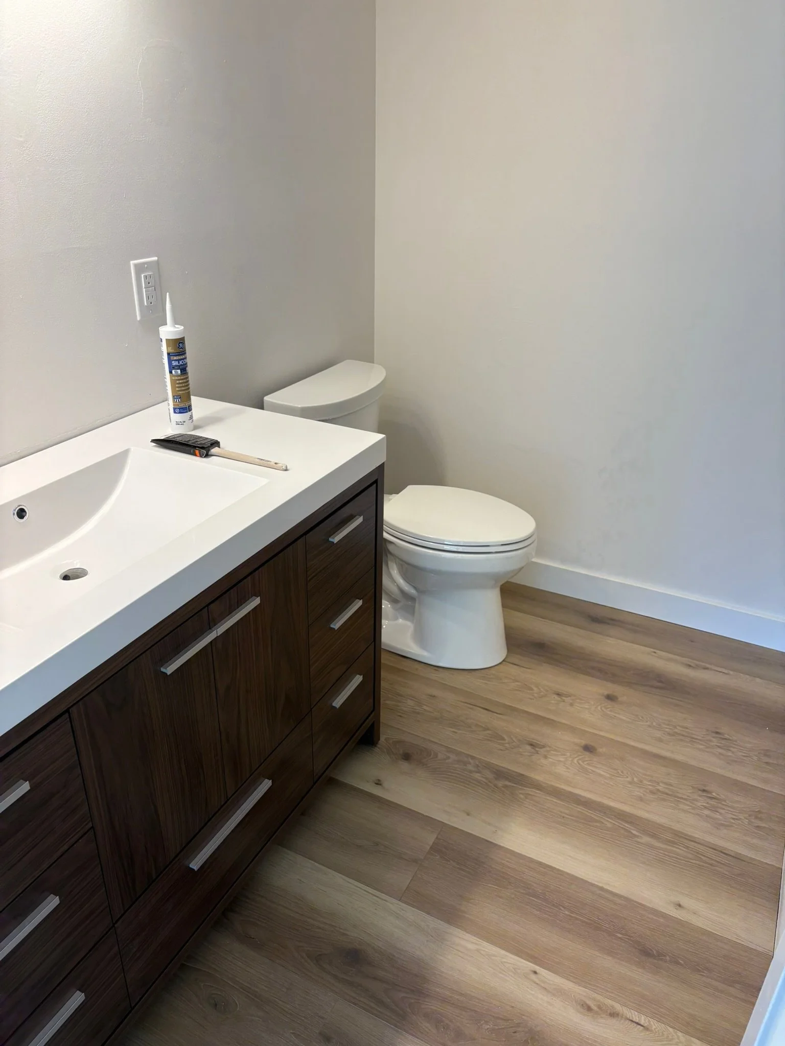 A bathroom with a white vanity and a wooden cabinet, a toilet, and a light wood floor. Items on the vanity include a tube of silicone, a tool, and a remote.