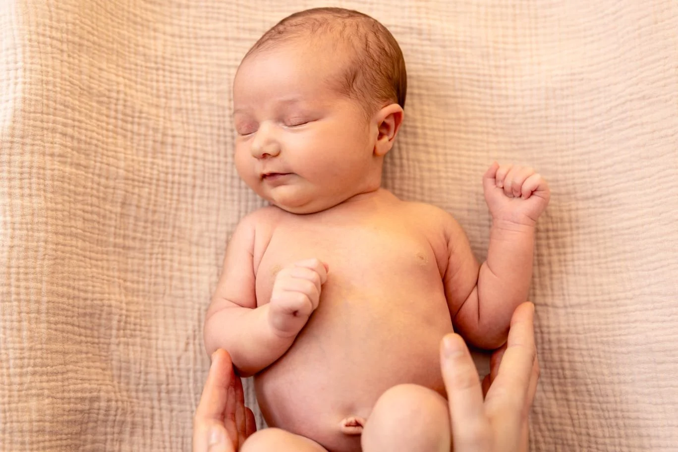 A sleeping newborn baby with closed eyes, on a soft beige blanket, with an adult hand gently holding the baby's arm.