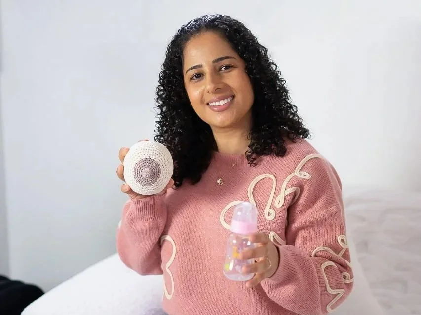 A woman with curly black hair smiling and holding a small round crocheted item in one hand and a pink baby bottle in the other, sitting on a white bed.