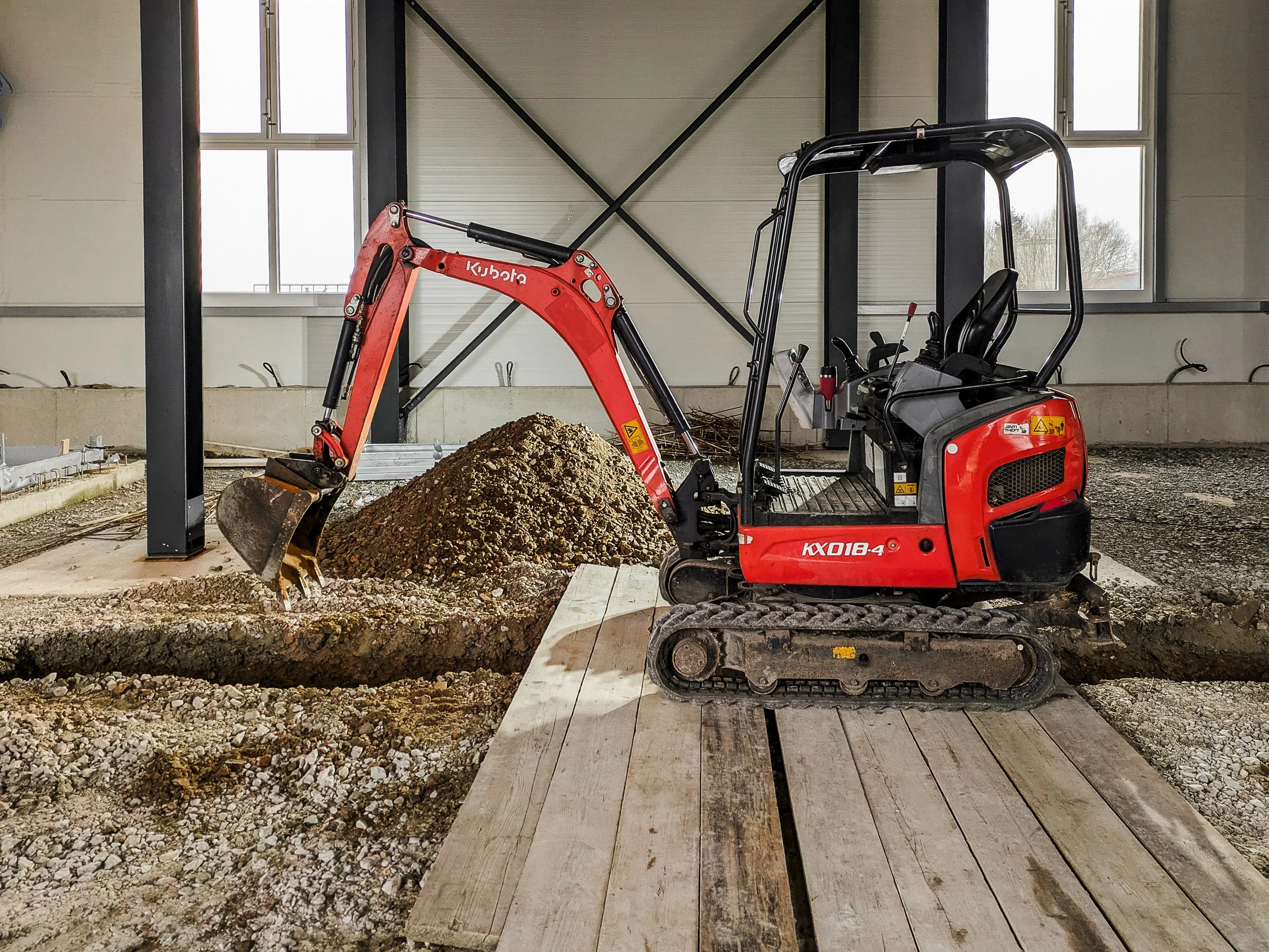 Kleines rotes Minibagger auf einer Baustelle, der einen Haufen Erde bewegt, im Hintergrund ein Gebäude mit großen Fenstern.