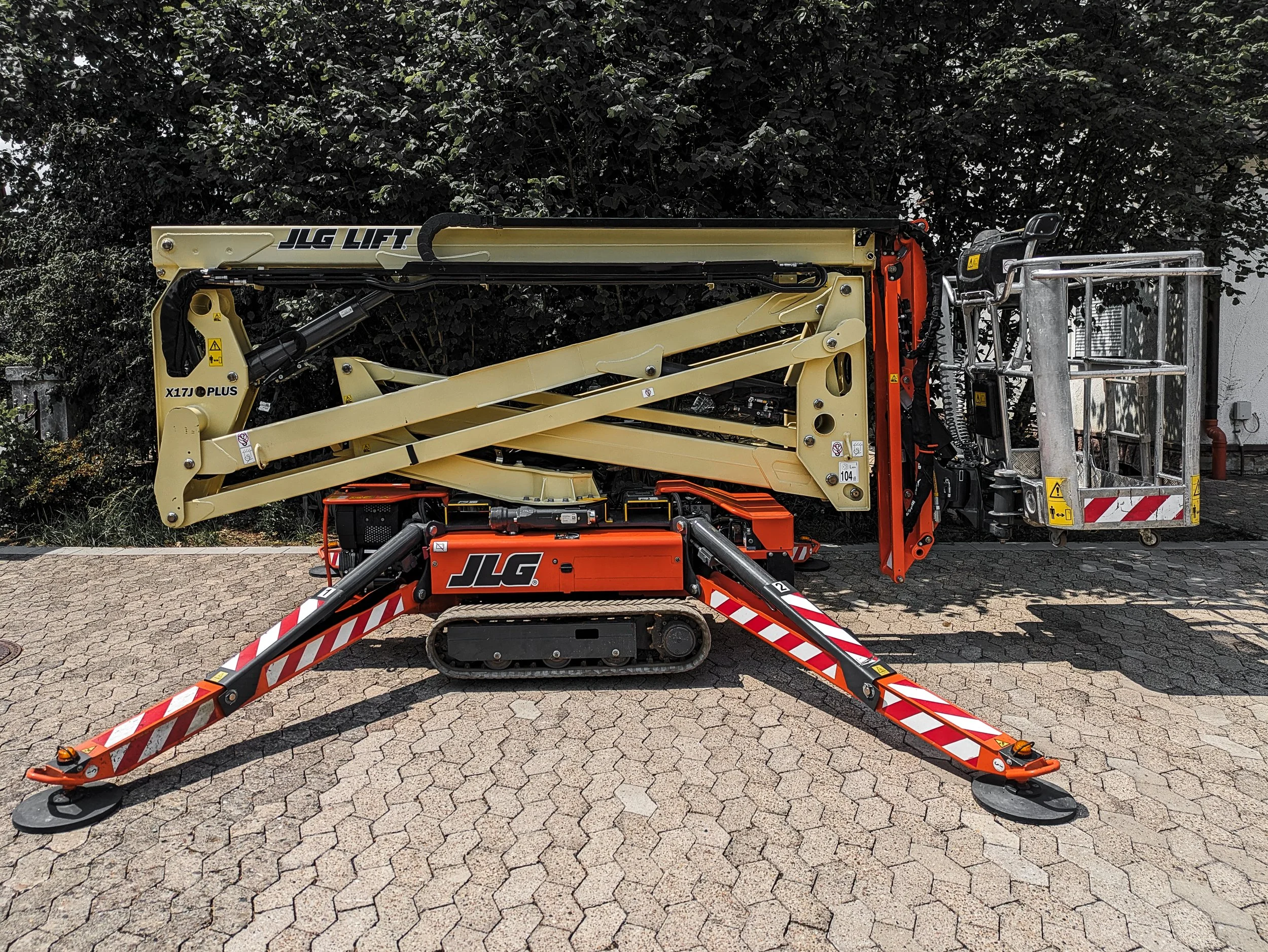 Ein gelber Hydrauliksteiger mit Korb, auf einem gepflasterten Boden, vor einer dunklen Baumhecke, mit rot-weißen Absperrbändern im Vordergrund.