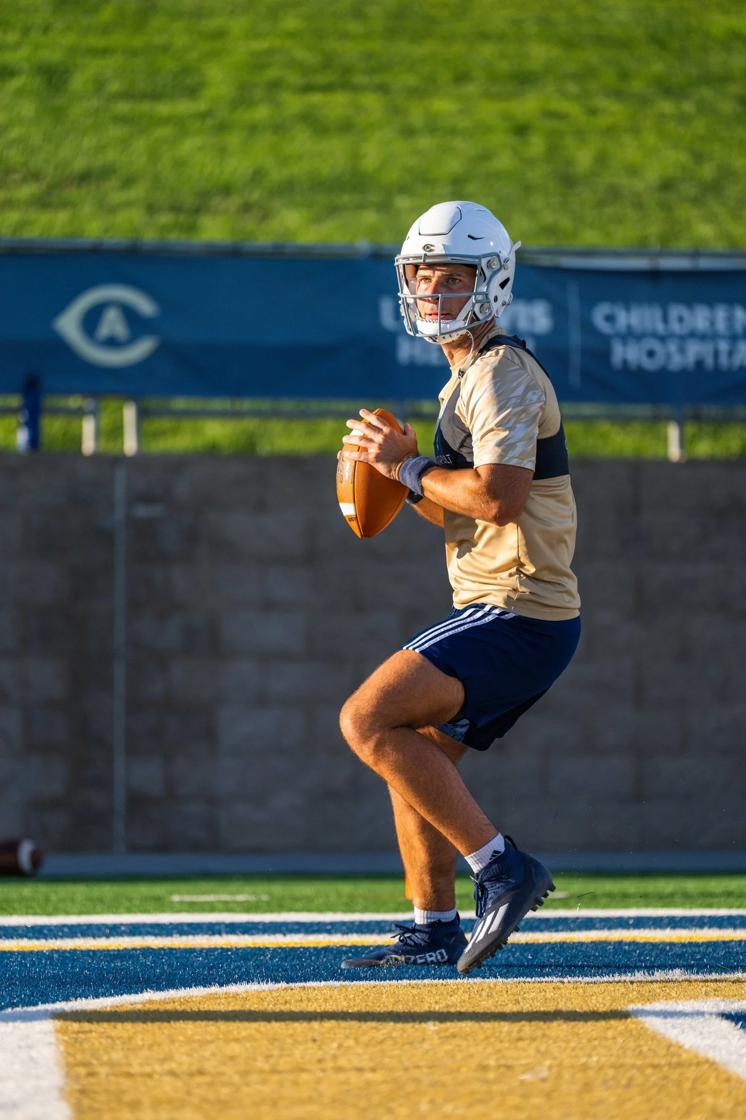 A man in football gear, including helmet and shorts, holds a football while standing on a field with colorful yard lines.