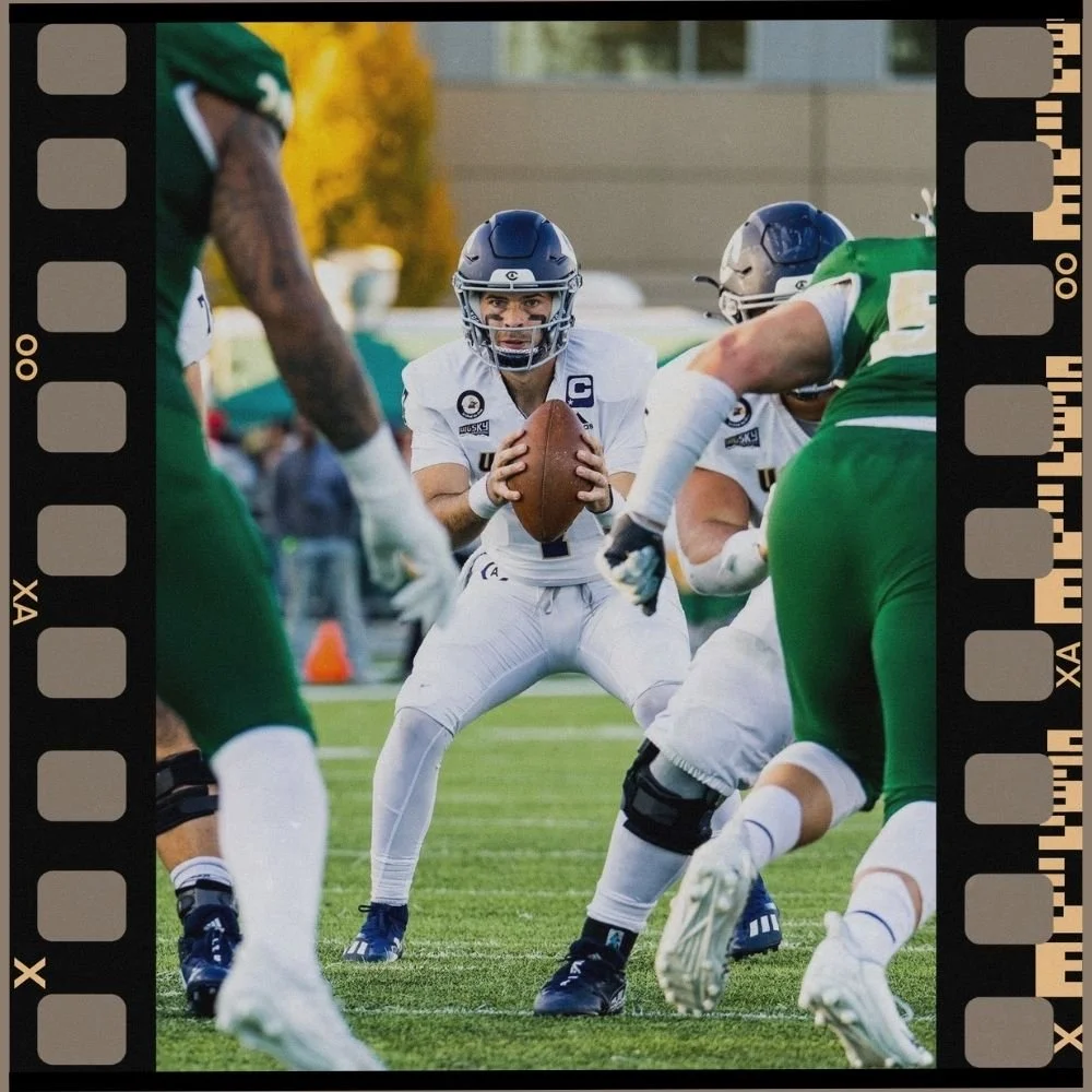 American football player in white uniform holding football, preparing to pass, facing a defensive line in green uniforms on a football field.