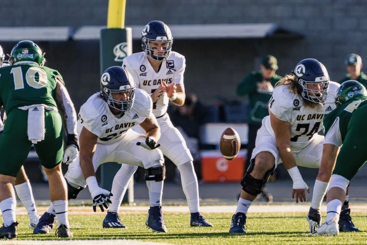Football players from UC Davis in white uniforms and a player from the opposing team in green uniforms preparing for a play on the field.