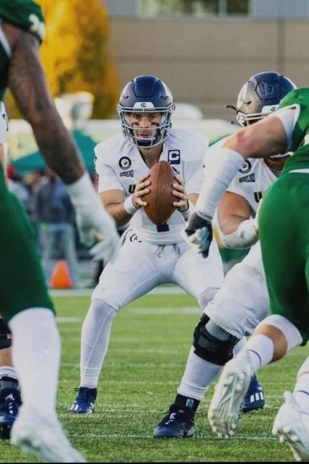 Football quarterback in white uniform preparing to throw the ball during a game, surrounded by players in green and white jerseys.