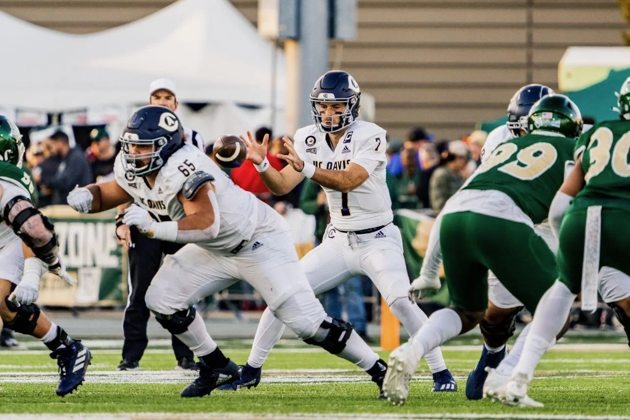 Football game with players in white and green uniforms, quarterback in white prepares to throw the ball, offensive line blocks, defensive players move against them on field.