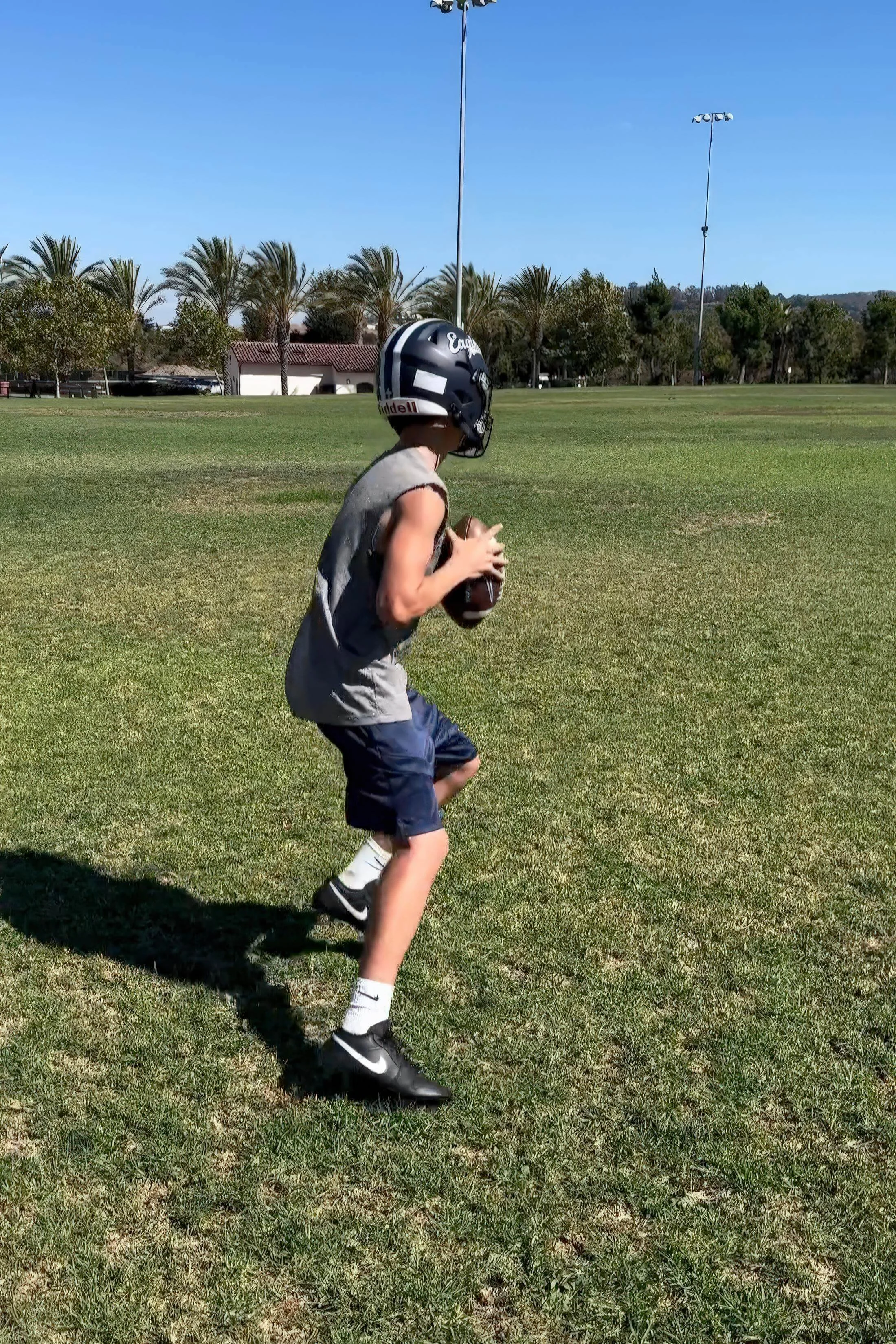 A young boy in a football helmet and athletic clothing running on a grassy field, holding a football, under a clear blue sky.