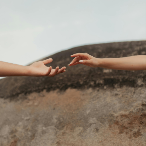 Two hands reaching towards each other against a rocky background and cloudy sky.